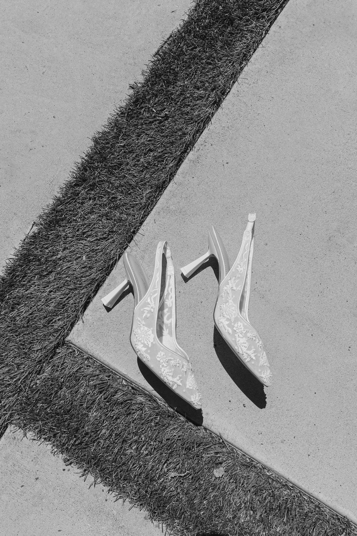 White lace wedding heels placed on concrete edge with grass strip in black and white.