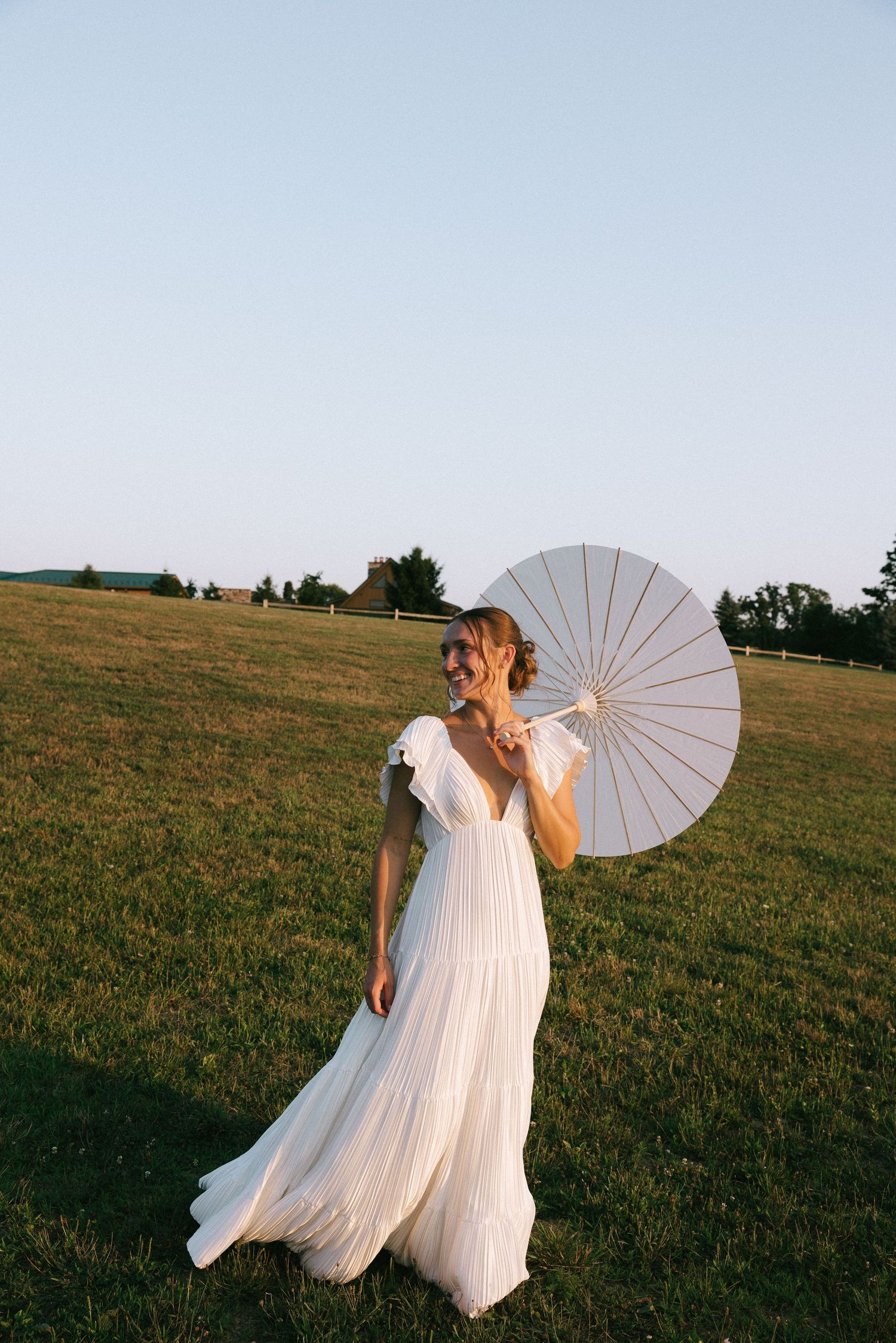 A bridal portrait in a flowing white dress holding a white parasol in a sunny meadow.