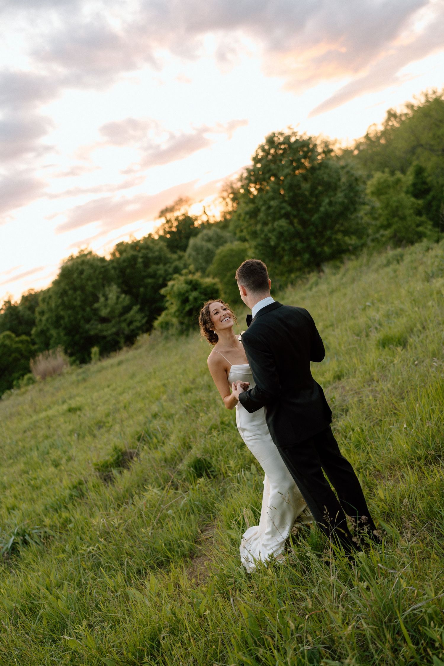 A romantic wedding couple sits together on a grassy hillside during golden hour sunset.