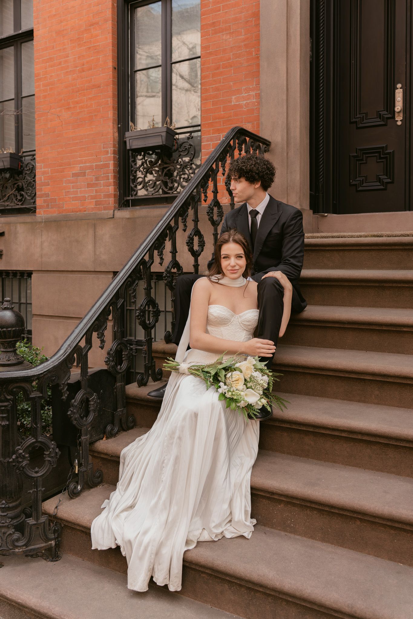 A couple in wedding attire sits on brownstone steps in an urban setting.