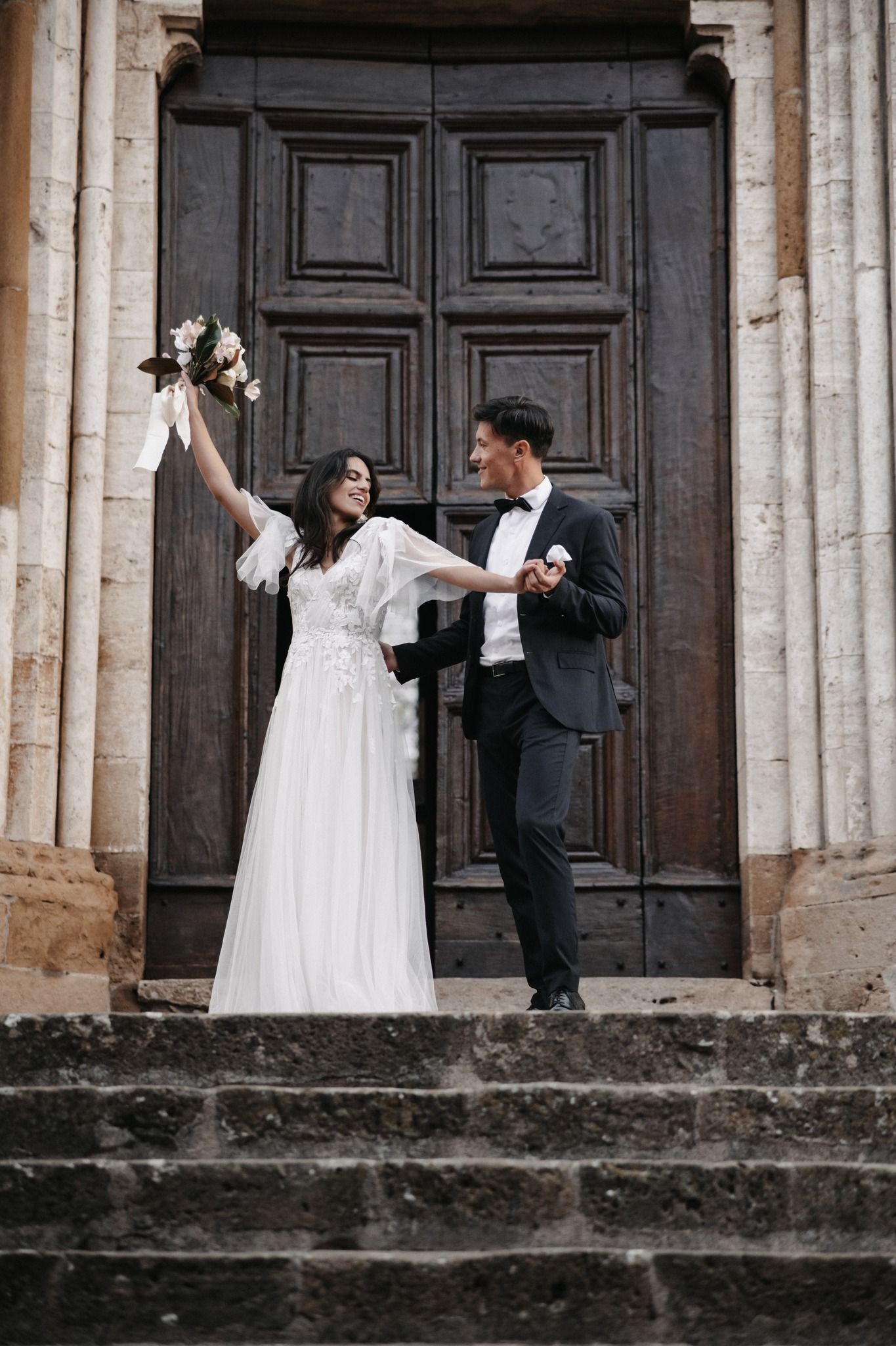 A celebratory moment on church steps as couple poses in front of large wooden doors.