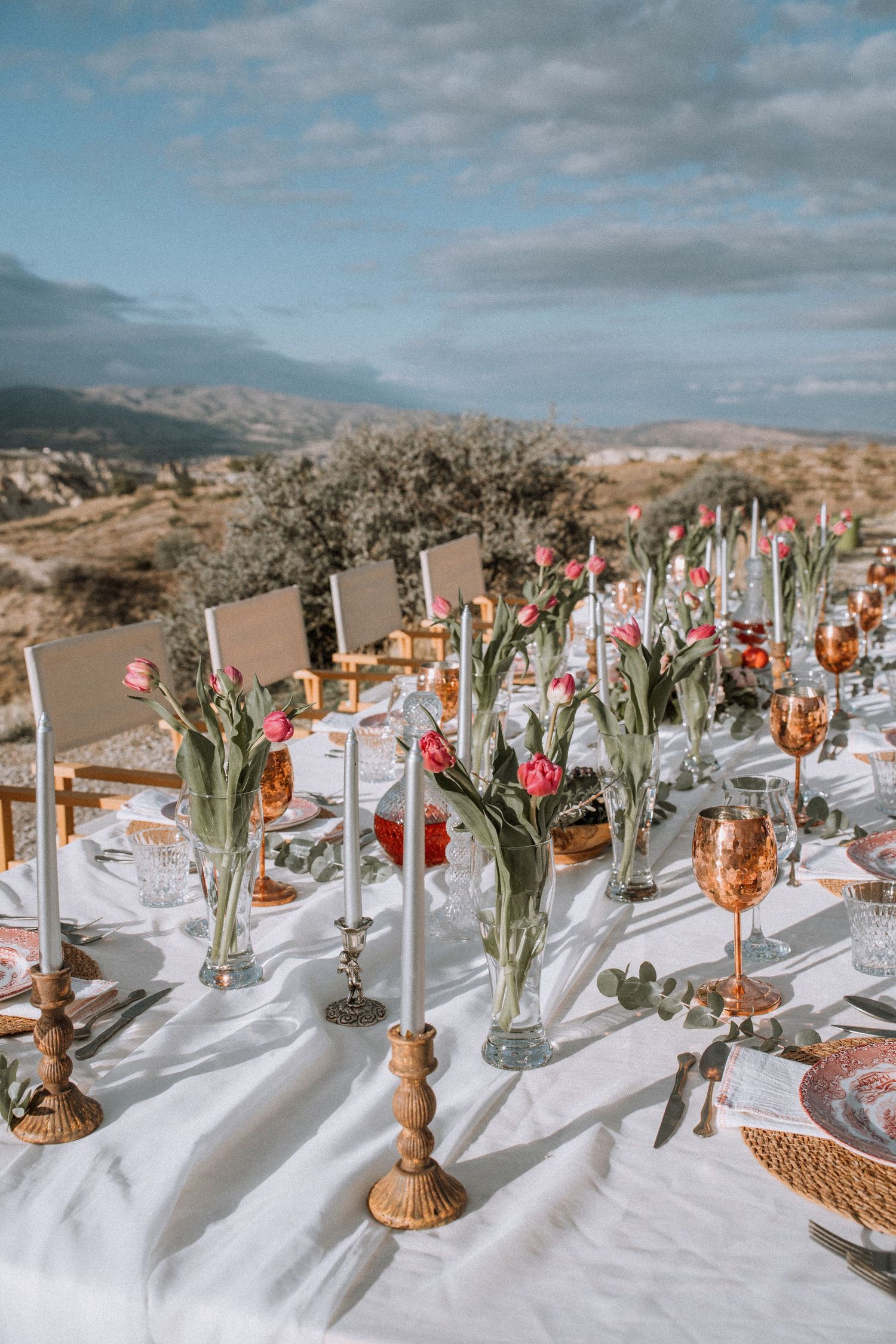 An outdoor wedding reception table setting with floral centerpieces against a mountain landscape.