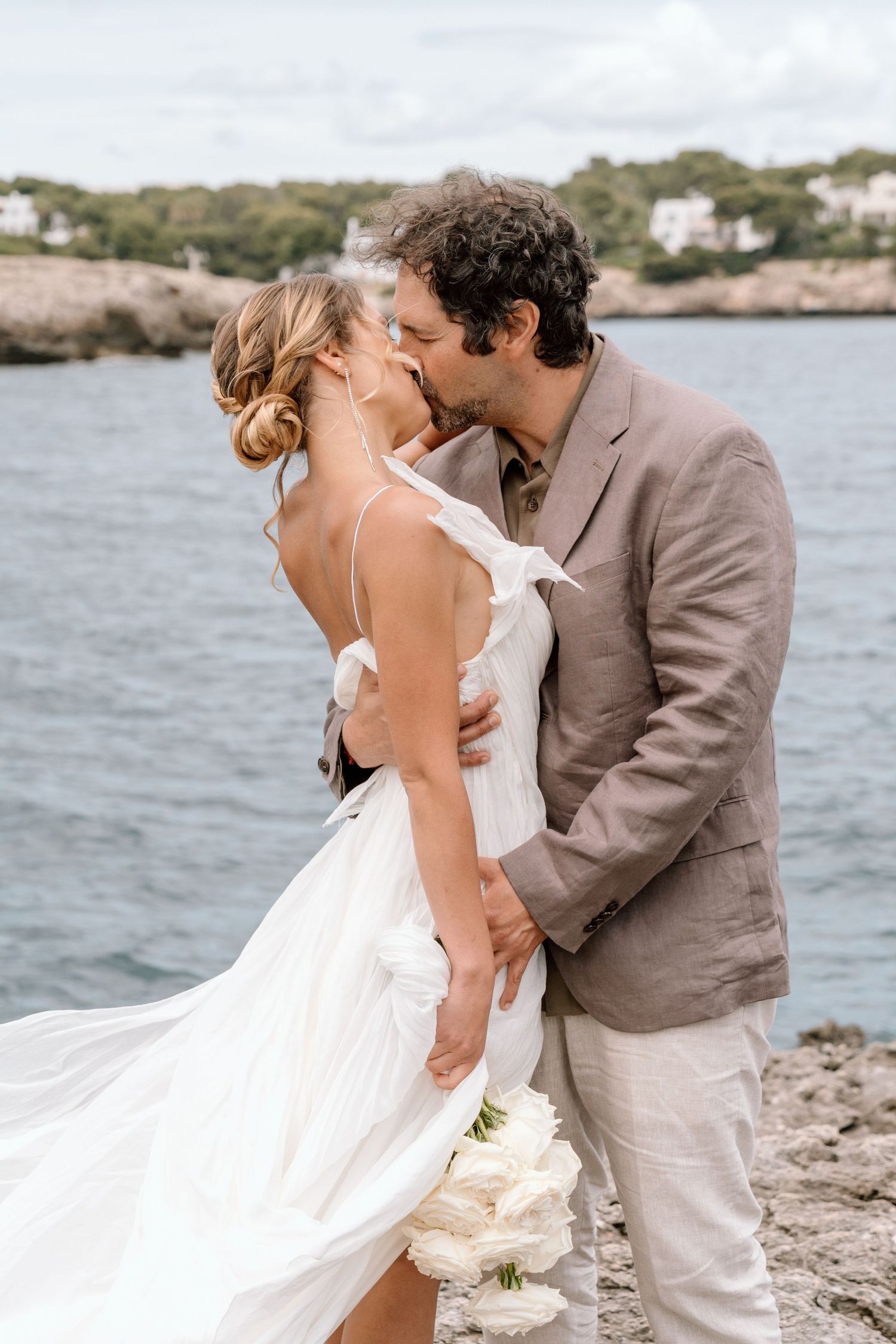 Wedding couple embracing on rocky coastal shore with white houses and blue water in background.
