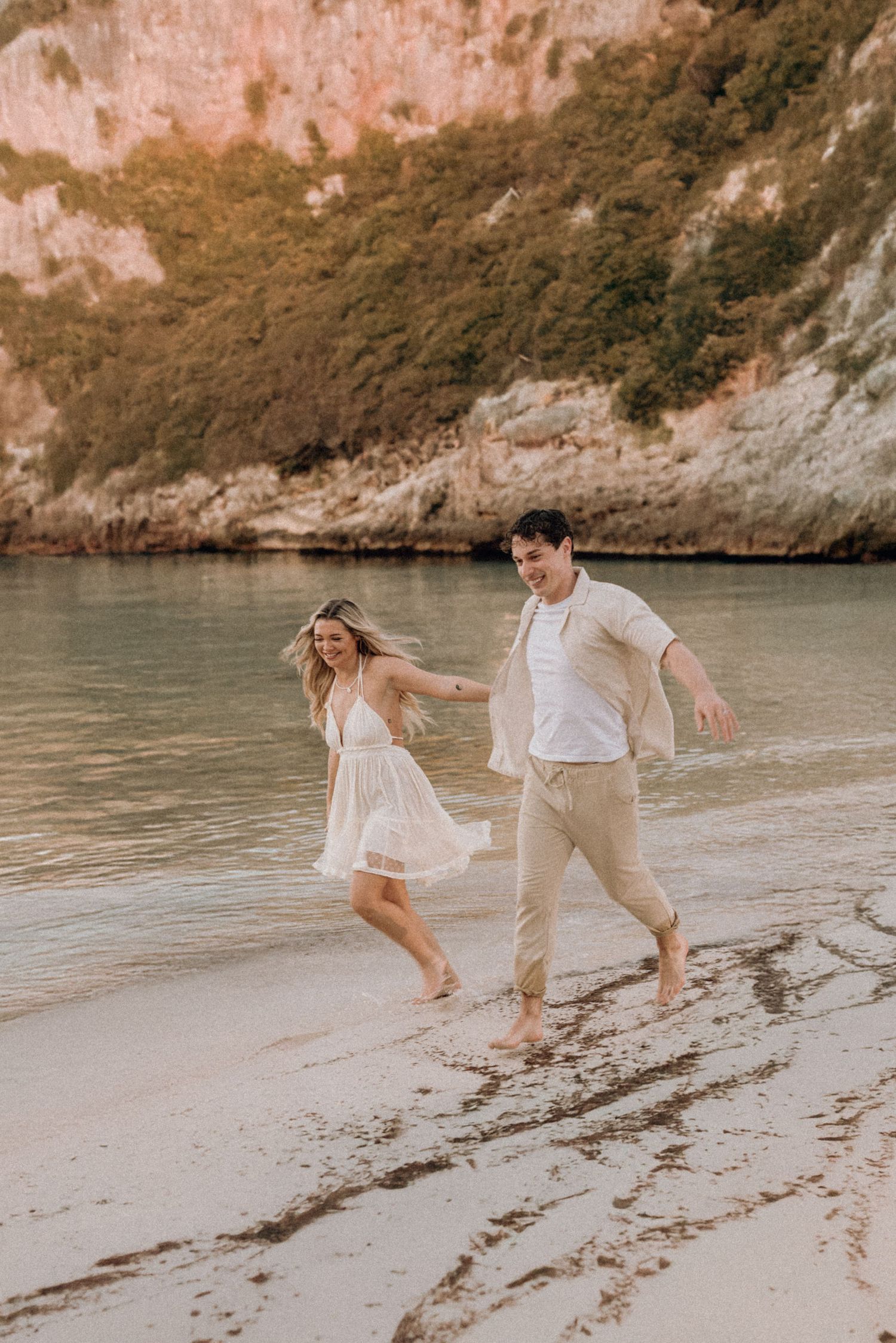 Playful couple in white outfits running and splashing along a beach at sunset.