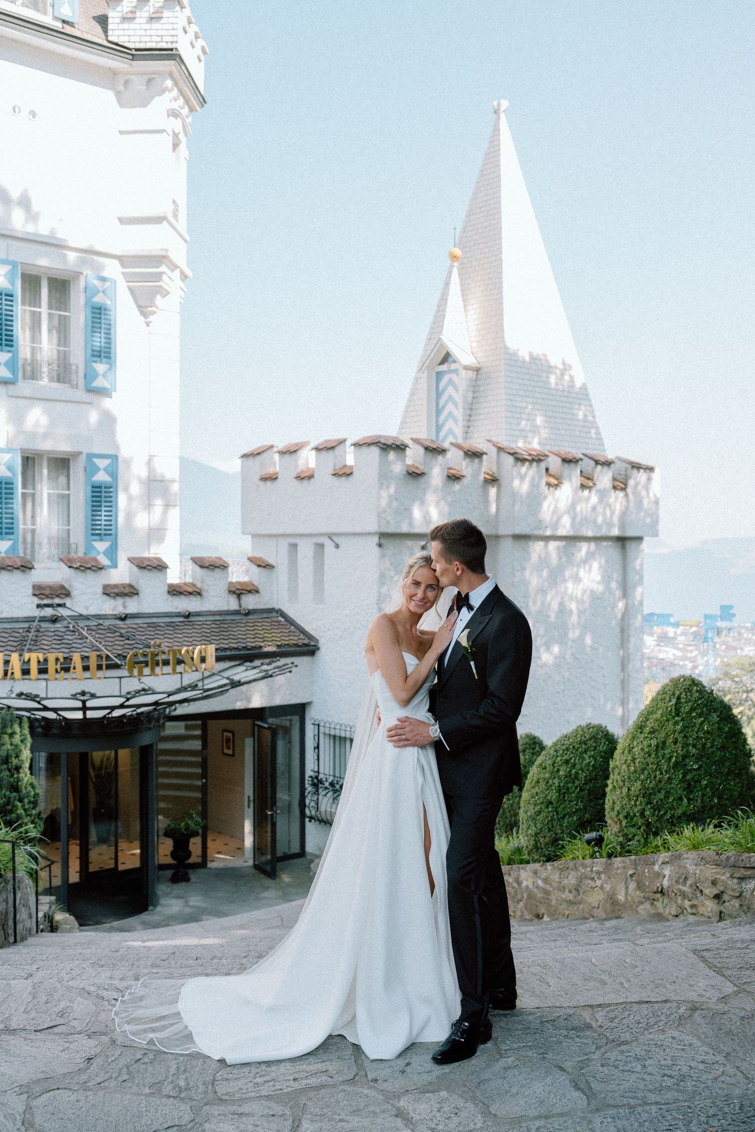 A romantic wedding couple embraces in front of a white castle spire with blue skies and mountains in the background.