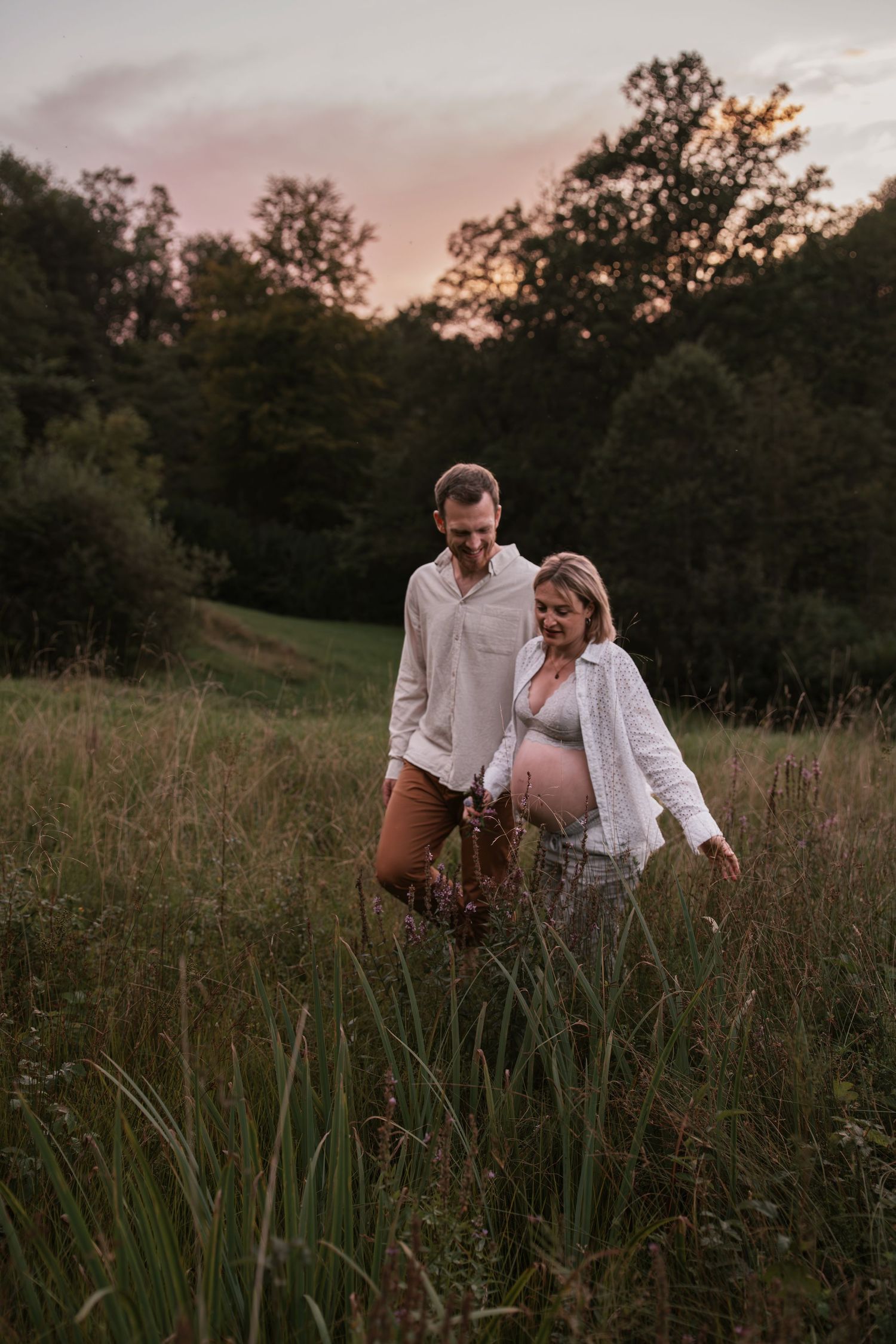 Couple walking through tall grass at sunset in a rustic field.