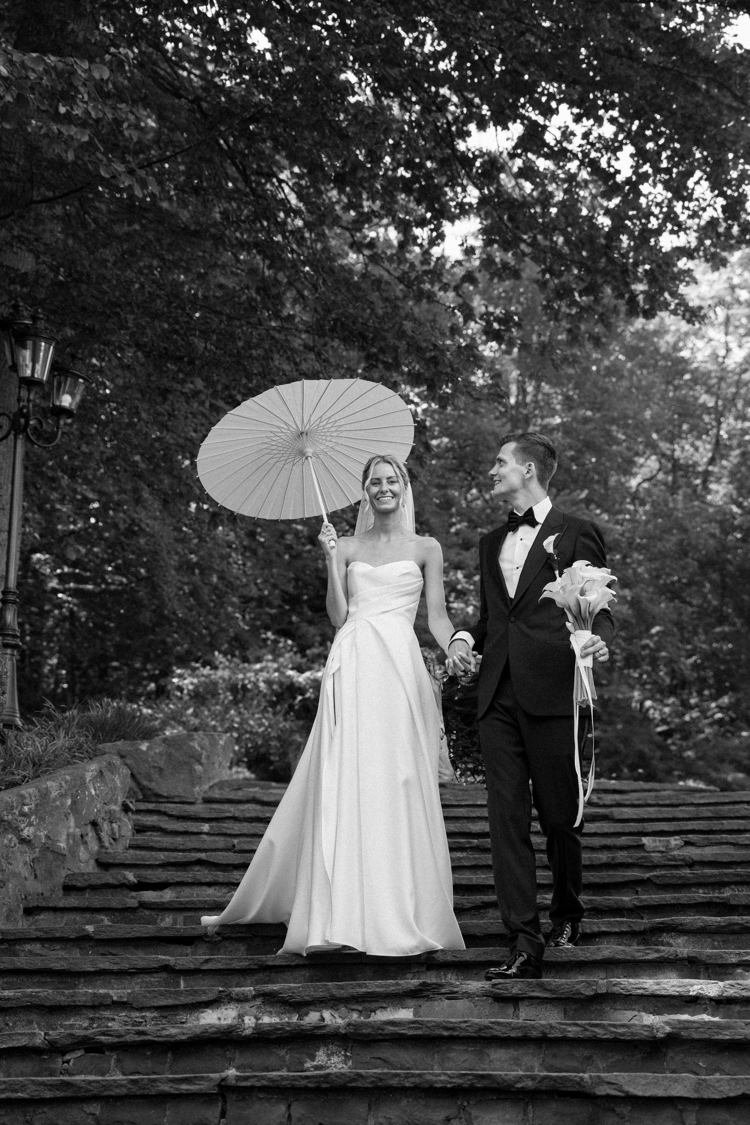 Wedding couple posing with parasols on stone steps beneath trees.