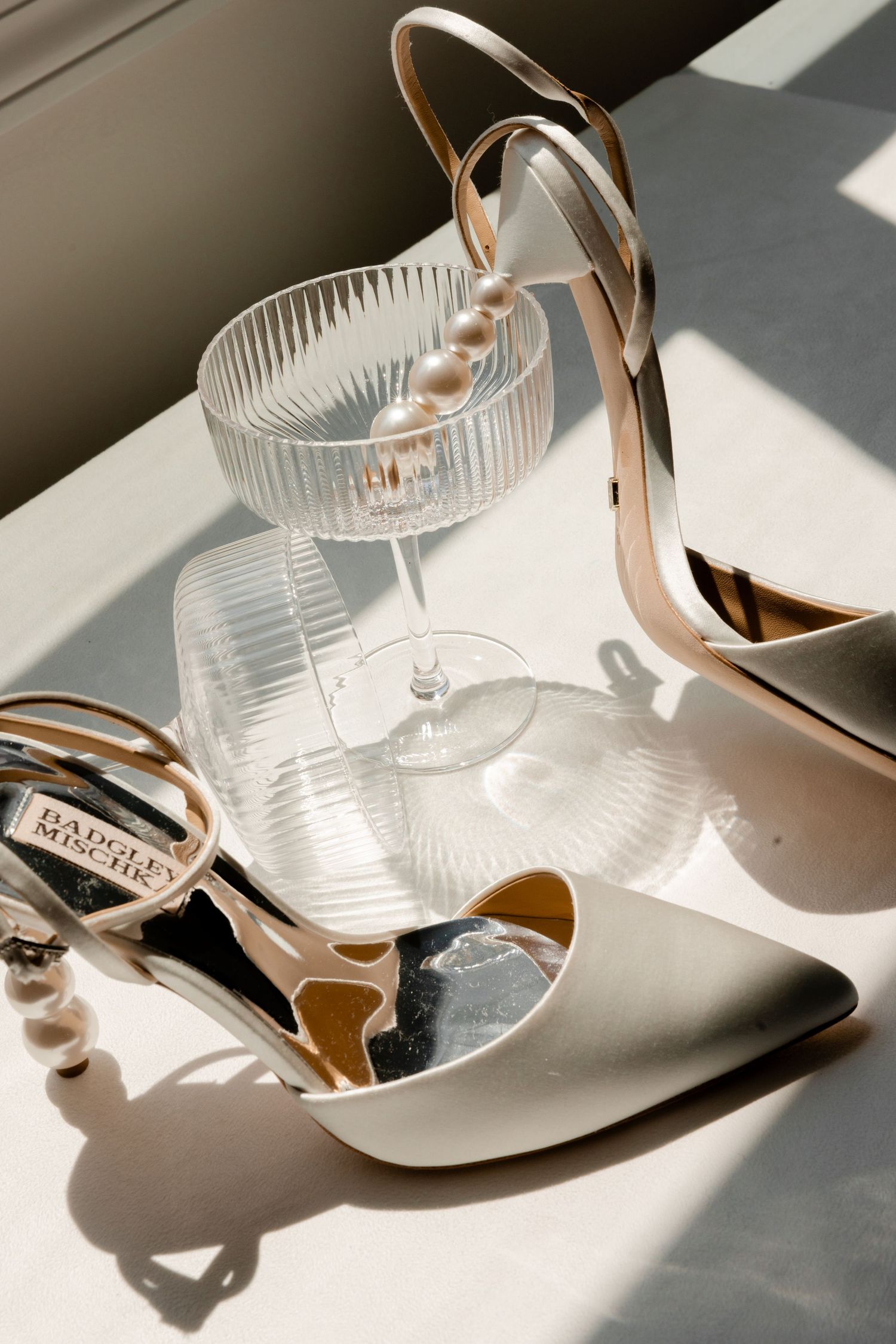 Elegant glass cocktail vessels and rose gold high heels arranged in artistic sunlit composition.
