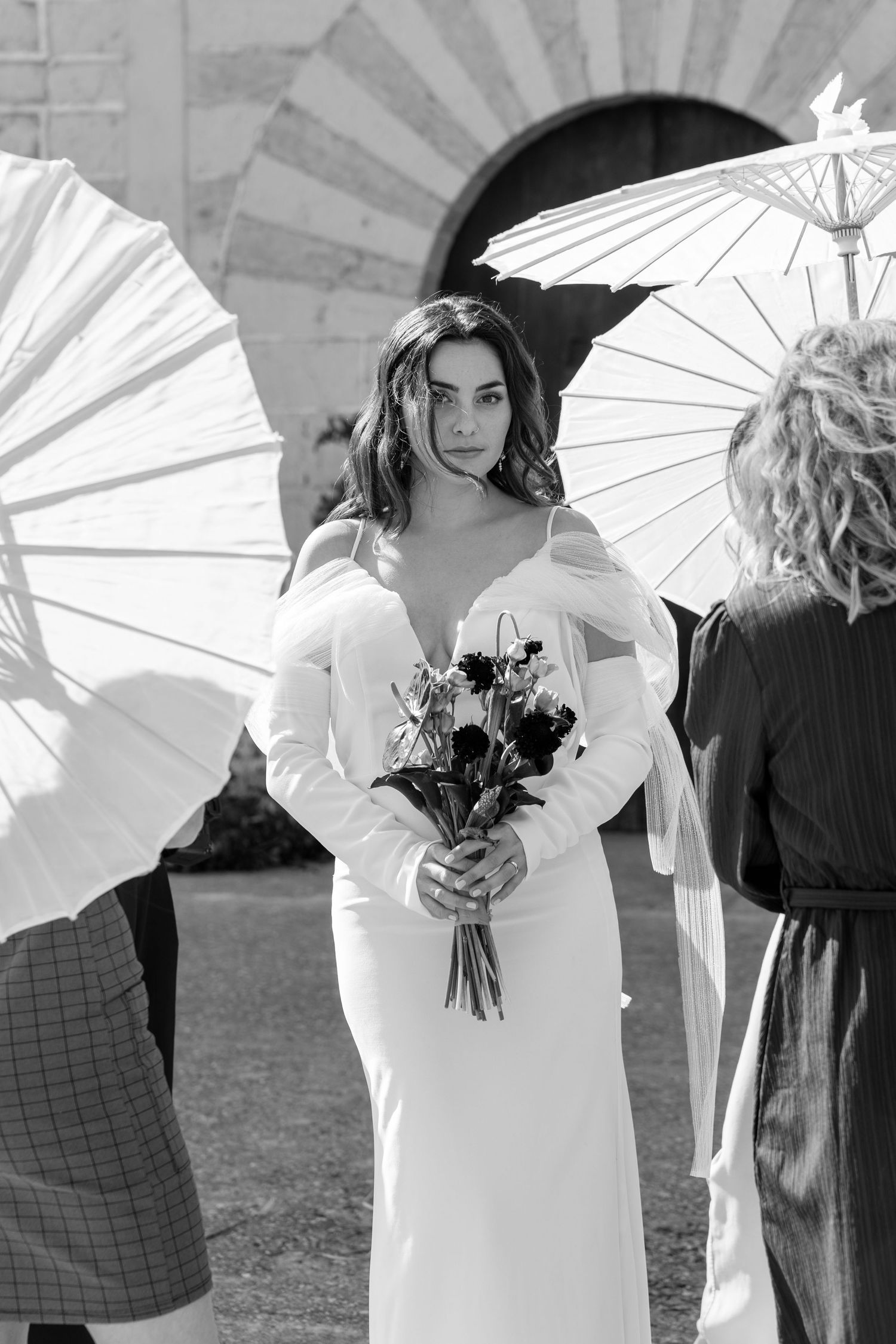 Wedding party holding white parasols against architectural backdrop.