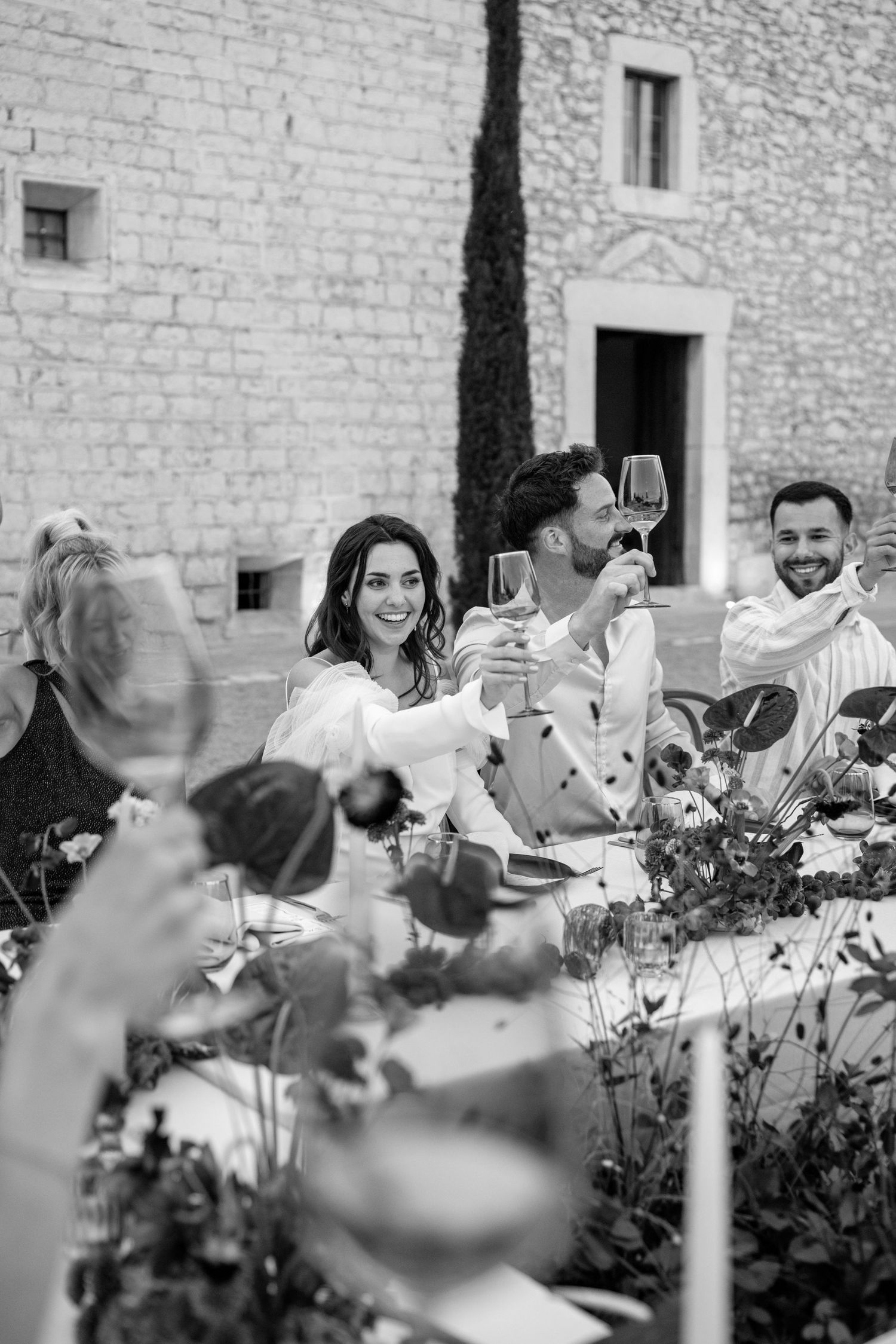Wedding reception scene with people gathered at tables near a stone wall.