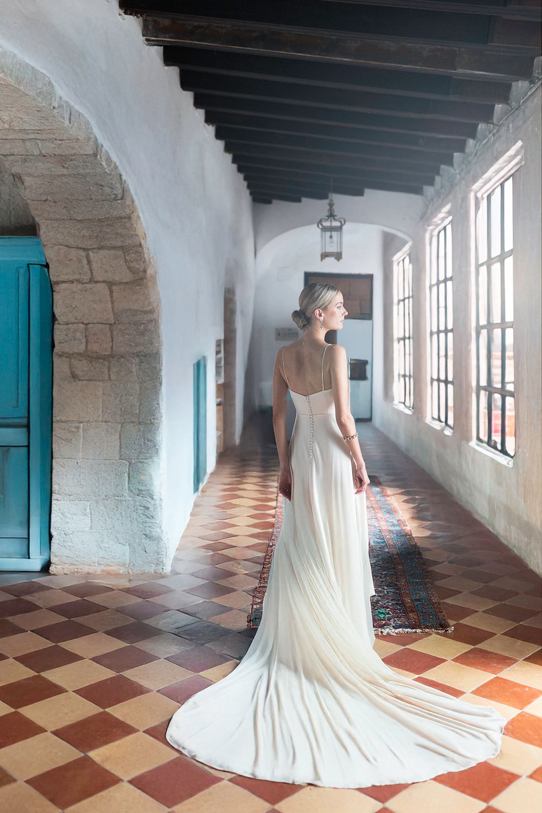 Bride in flowing white gown standing in Spanish colonial corridor with checkerboard floor and arched windows.
