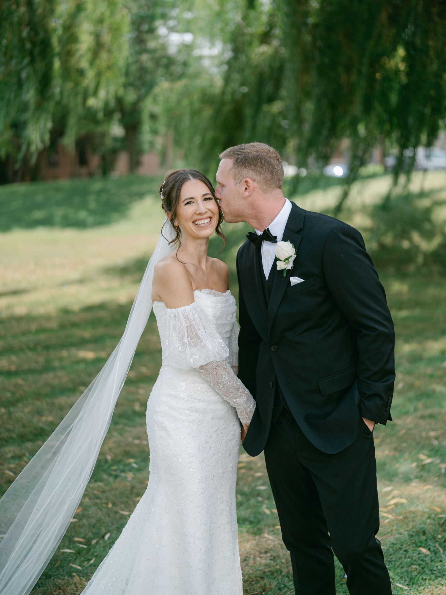 A romantic wedding couple shares intimate moments under willow trees in an outdoor garden setting.