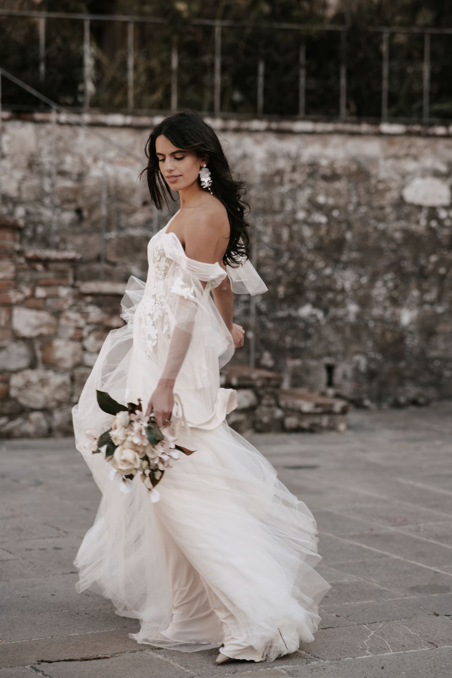 An elegant white wedding dress flows and twirls against a rustic stone wall backdrop.
