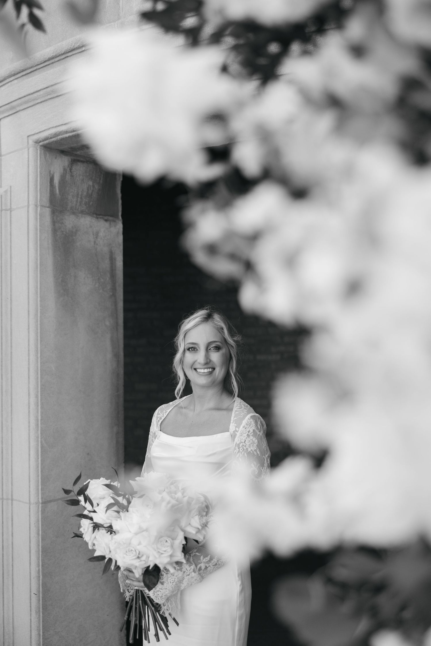 A monochrome bridal portrait featuring a flowing white dress and bouquet framed by delicate florals.