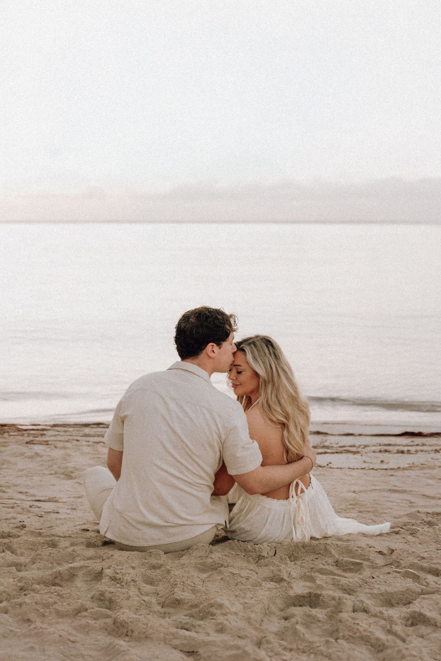 A romantic couple sits close together on a serene beach at sunset.