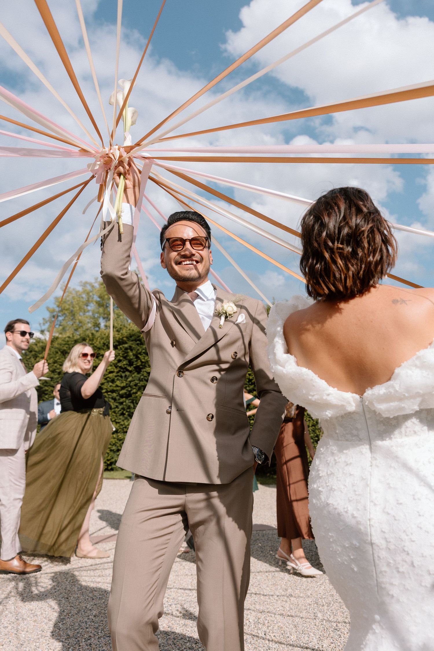 Wedding party celebrates under copper-spoke umbrella structure against blue sky.
