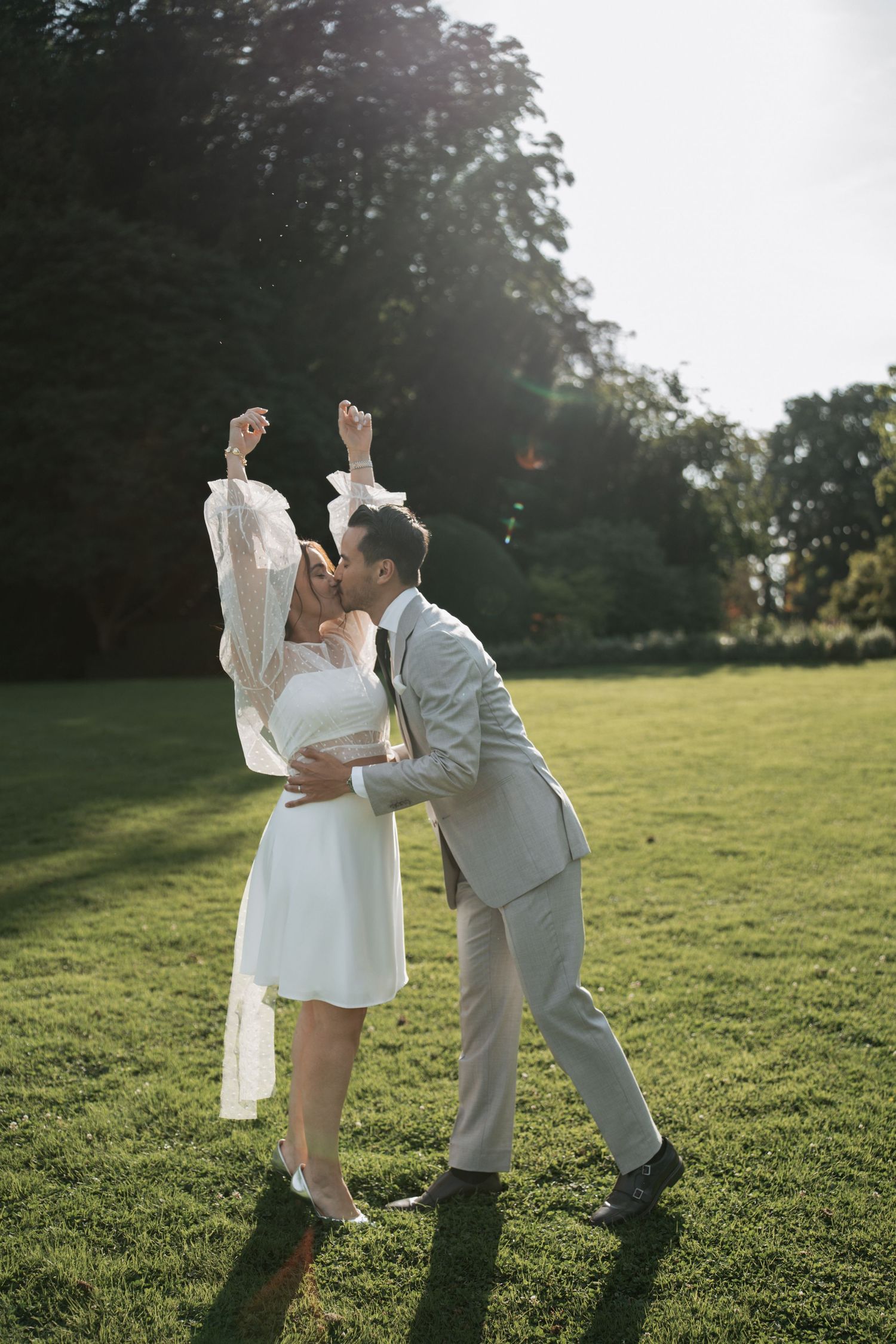 A wedding couple shares a playful moment on a sunny grass lawn.