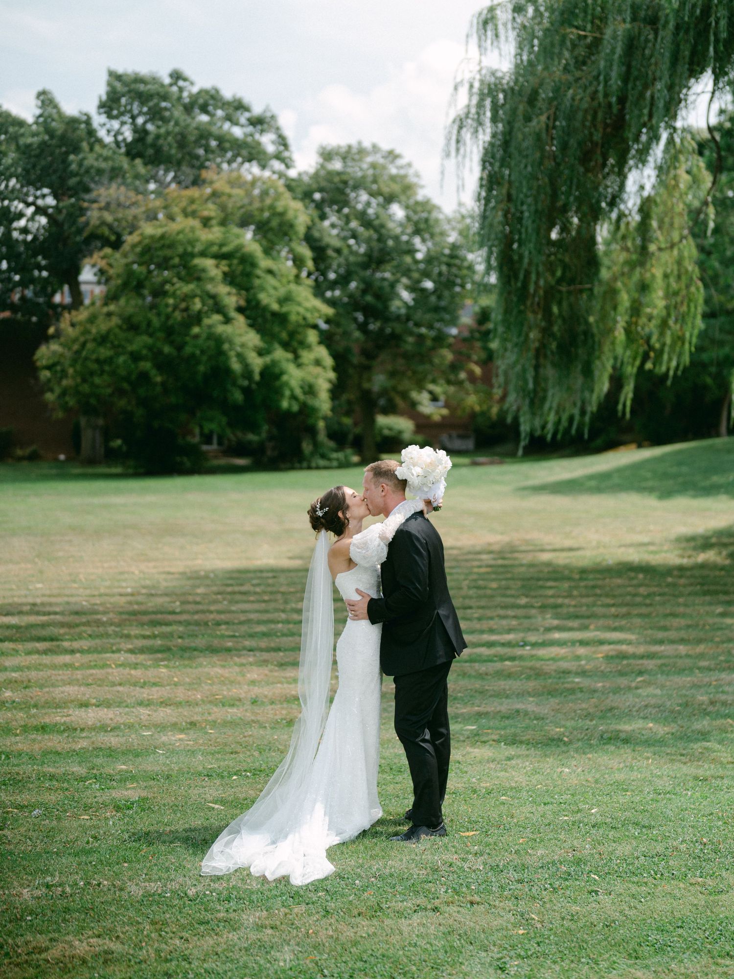 Romantic wedding couple embracing on lawn with weeping willow trees in background.