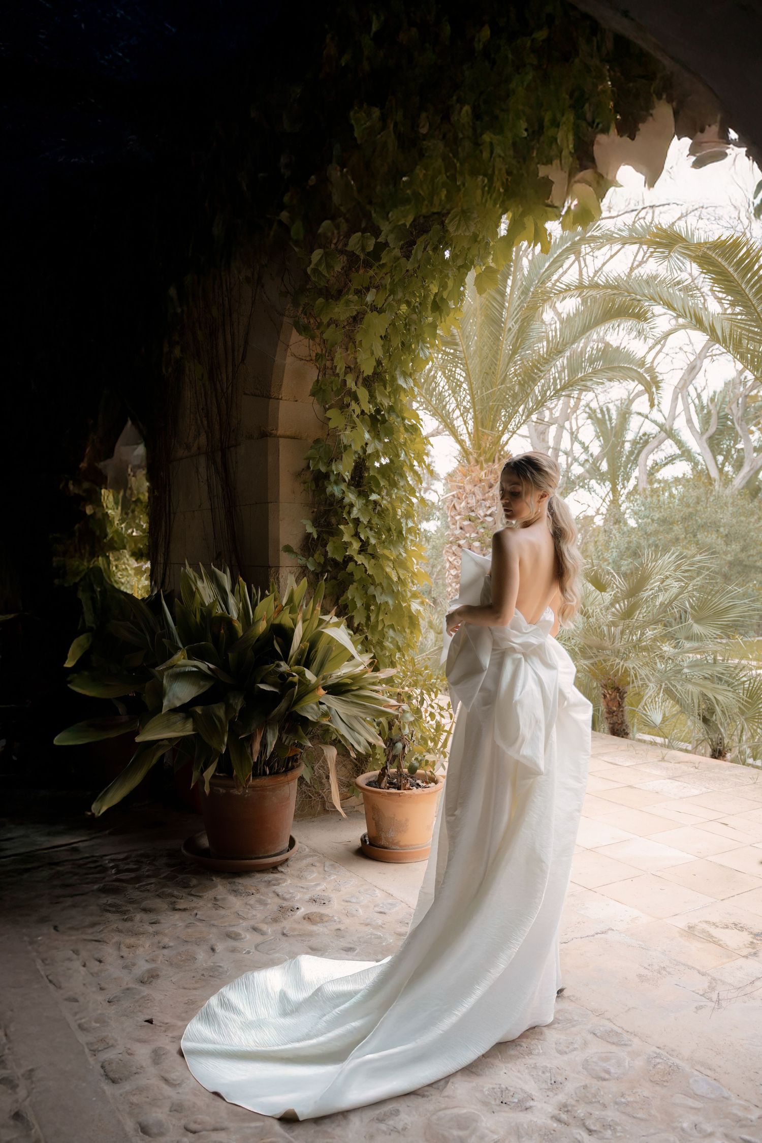 Bride in flowing white dress photographed from behind on Mediterranean-style terrace with palm trees.