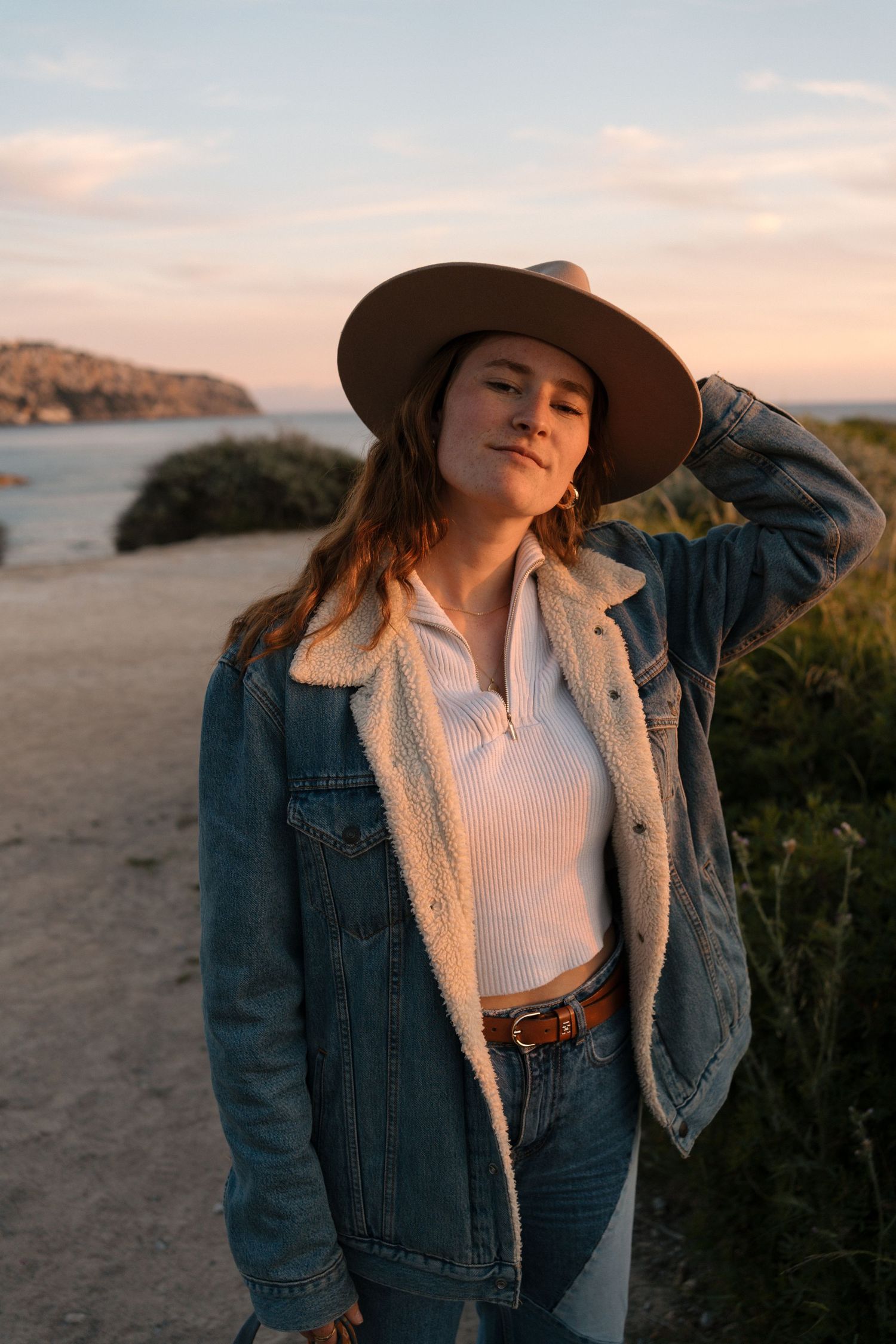 Person in denim jacket and wide-brimmed hat poses against coastal sunset backdrop.