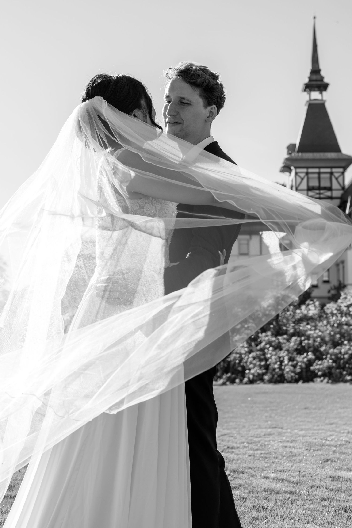 Black and white wedding photo of a bride and groom embracing on a lawn as her sheer veil billows in the wind near a historic tower.