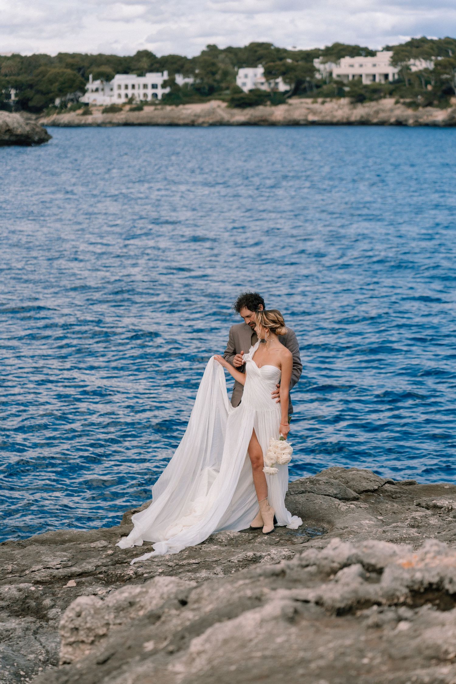 Wedding couple sitting on coastal rocks overlooking blue Mediterranean waters with white dress flowing.
