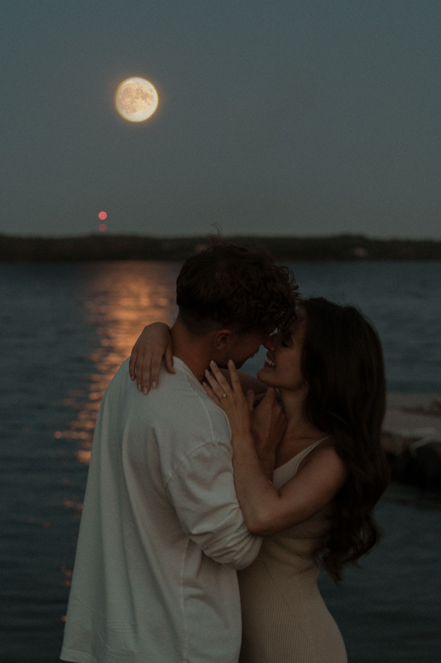 Couple sharing romantic kisses under full moon by moonlit water.