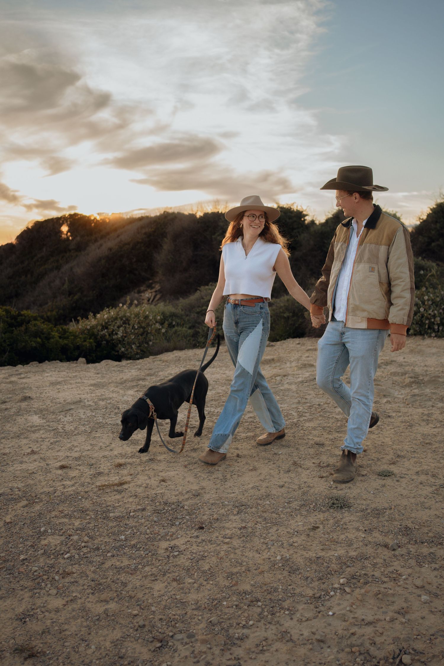 Couple walking with their dog during golden hour on a hilltop trail.