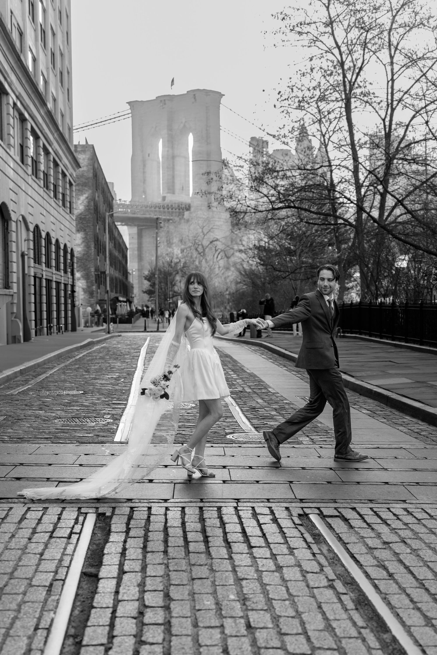 Series of playful wedding photos on cobblestone street with Brooklyn Bridge in background.