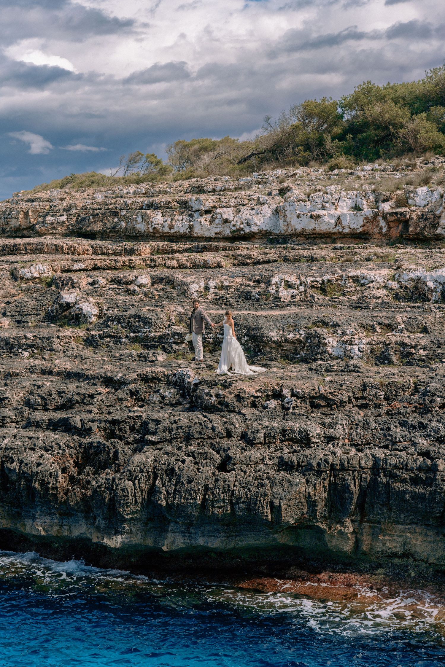 Rocky coastal cliffs overlooking deep blue Mediterranean waters with a small sailboat in the distance.
