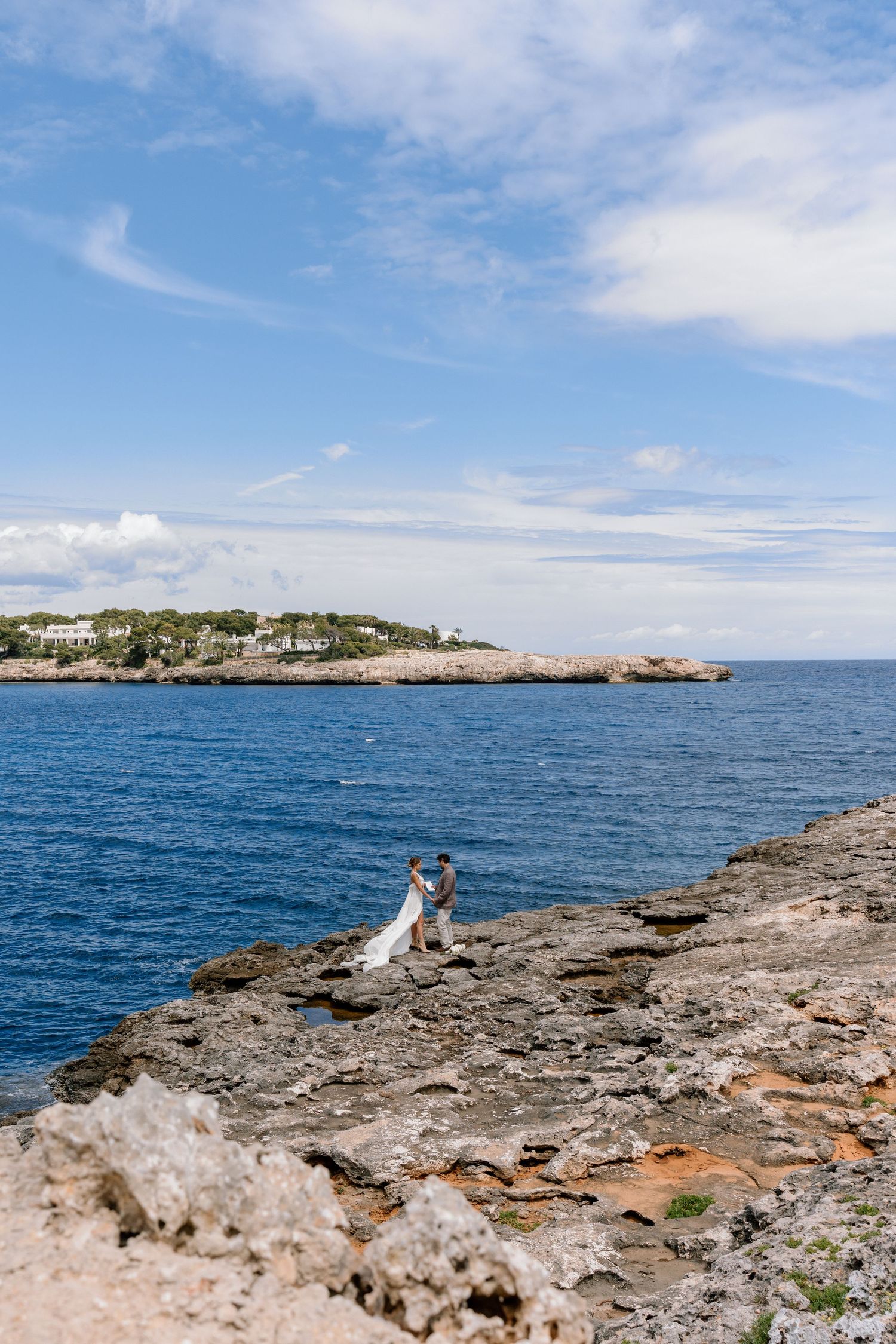 Wedding photography on rocky coastal cliffs with bright blue ocean waters and white seaside buildings in distance.