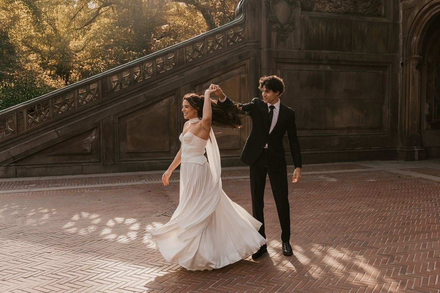 A wedding couple twirls together on a brick pathway with sunlit architecture in the background.