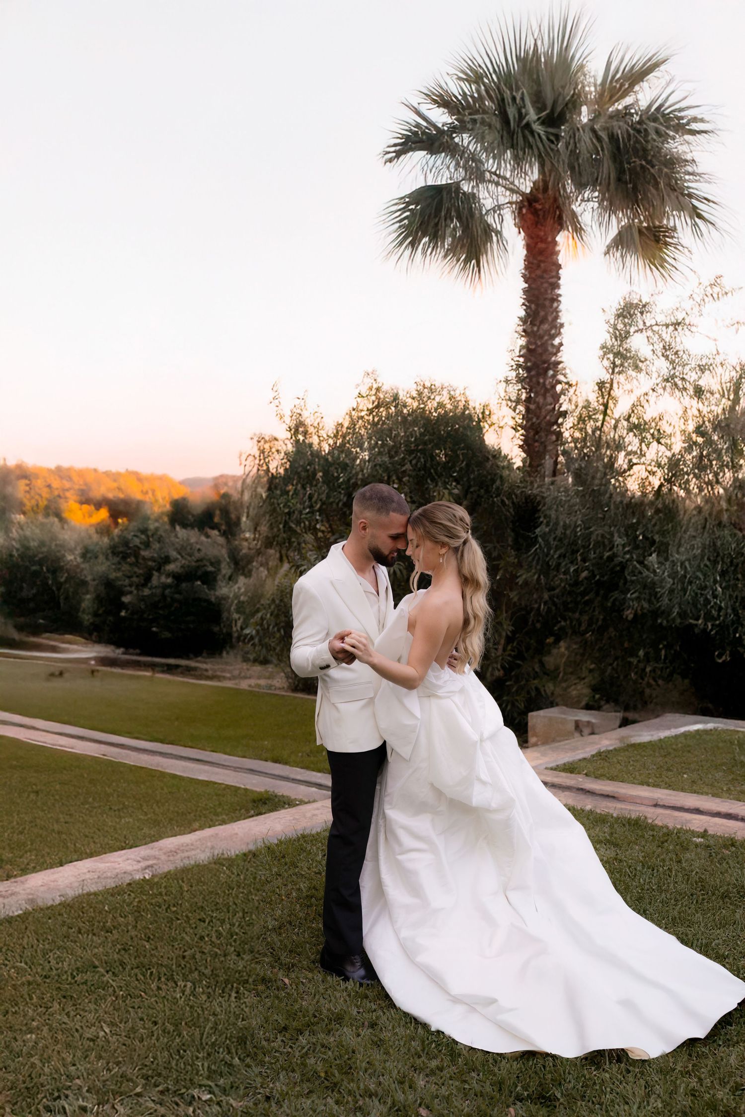 A sunset wedding photoshoot with palm trees shows a dancing couple in formal attire on a lawn.
