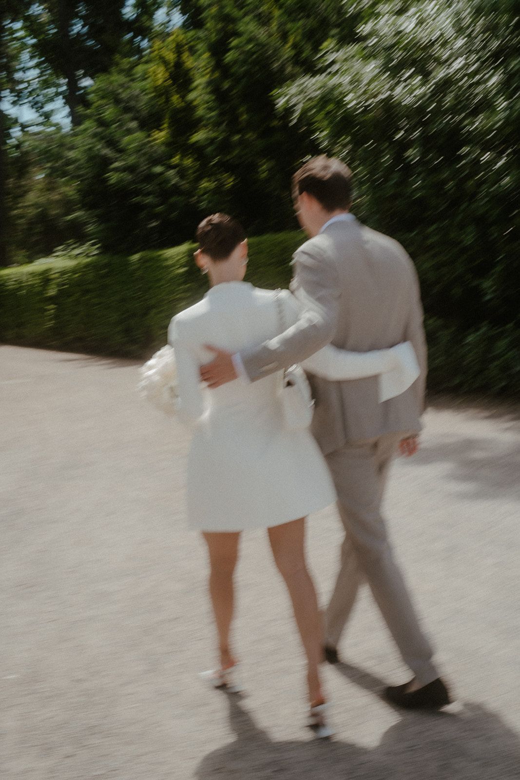 Couple in formal attire playfully walk and dance together on a sunlit garden path.