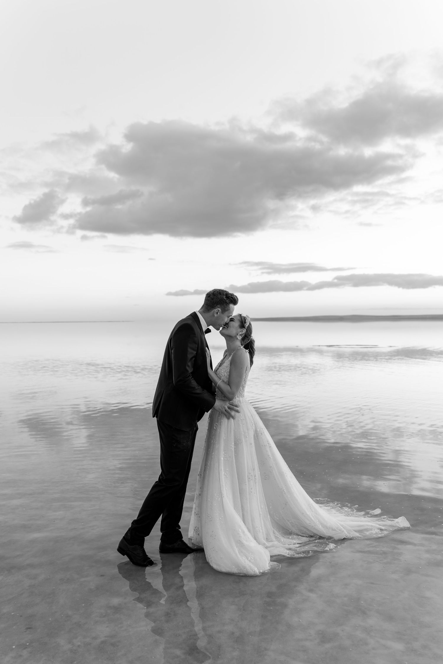 A couple shares an intimate moment on a beach at sunset with reflected light on wet sand.