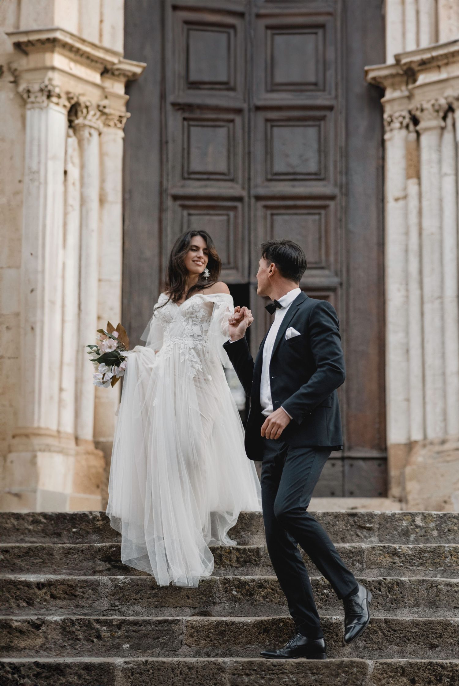 A couple descends stone cathedral steps, the bride in a flowing white gown and groom in classic black tuxedo.