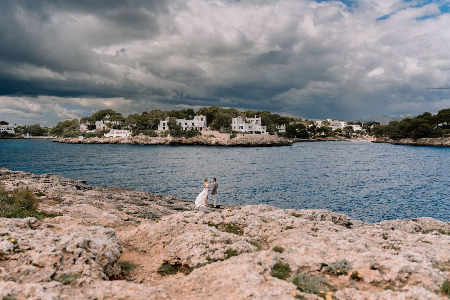 Panoramic coastal view with rocky shoreline and Mediterranean buildings under cloudy skies.