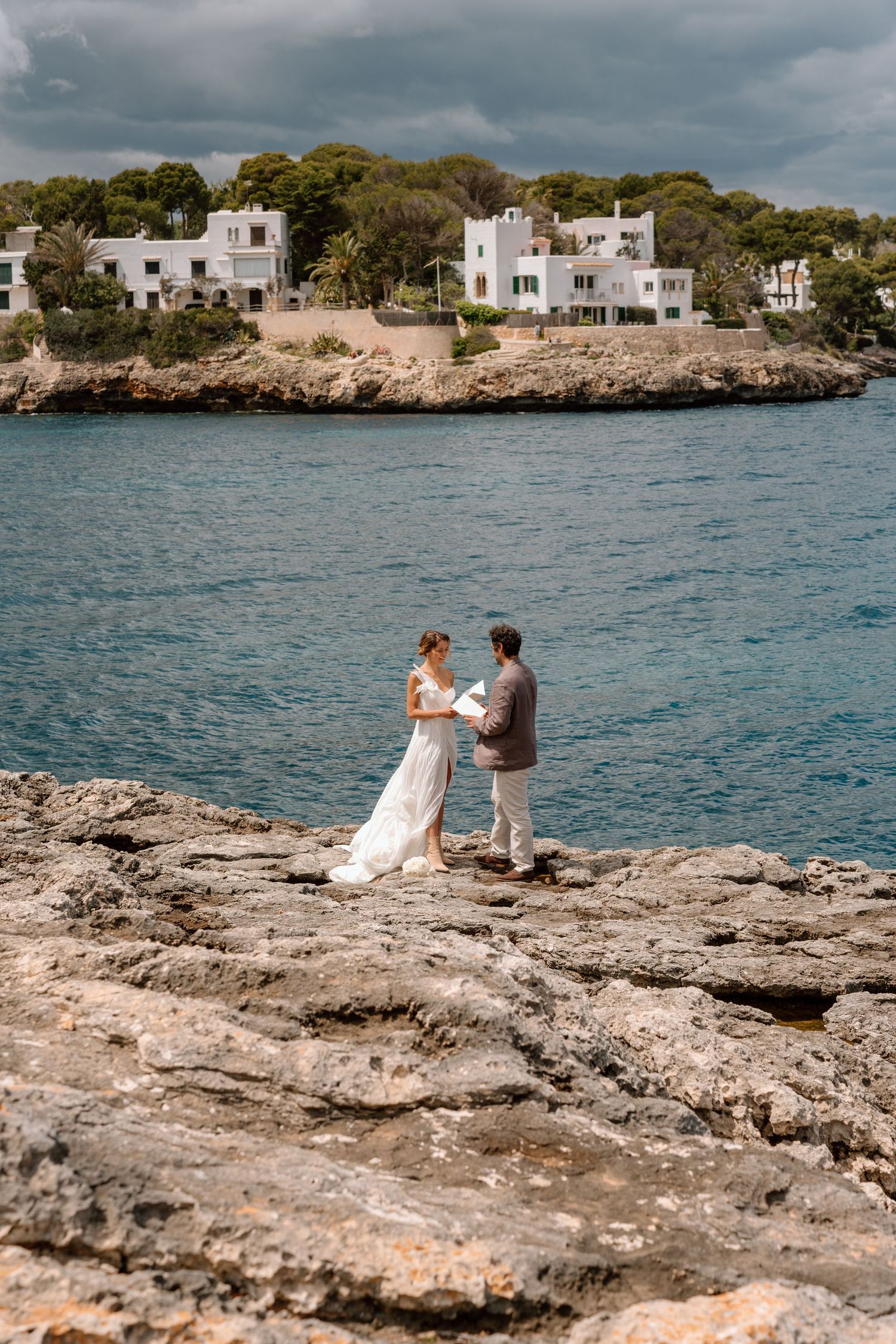 Wedding couple embracing on rocky Mediterranean coastline with white villas in the background.