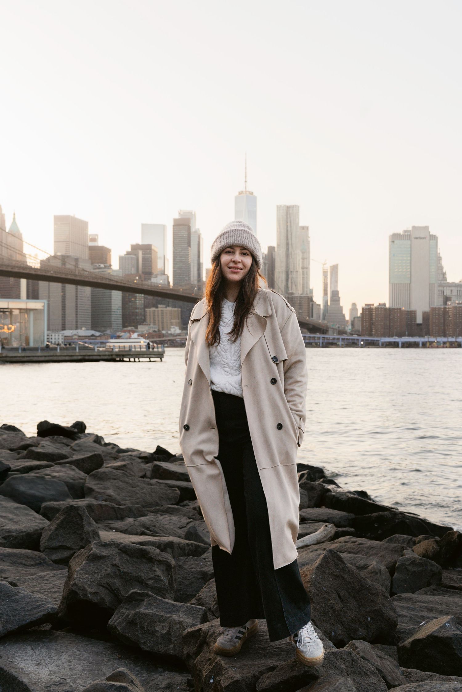 Person in beige coat stands along rocky shoreline with Manhattan skyline at sunset.