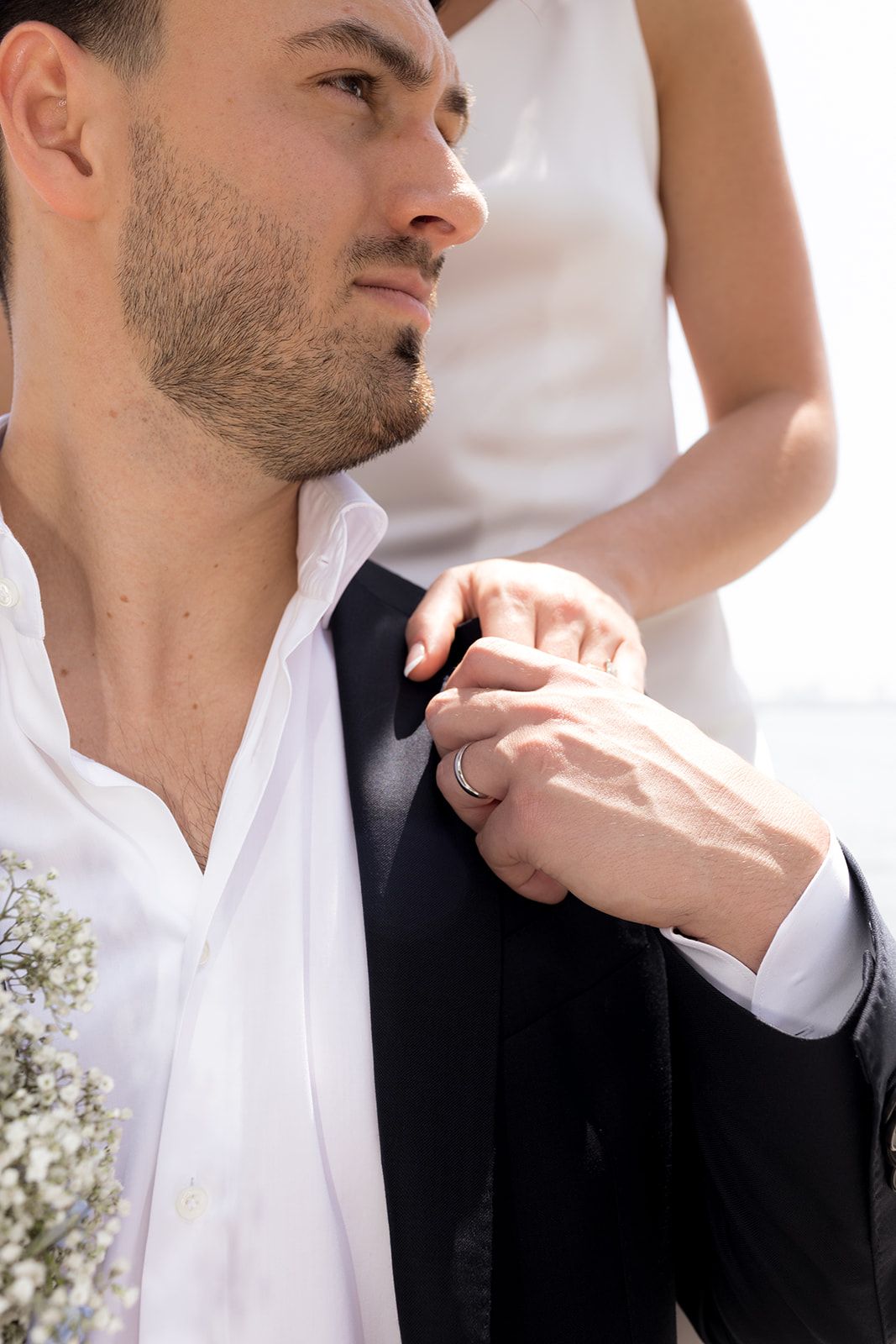 Bride's hand rests on groom's dark suit jacket shoulder as he gazes into the distance, wearing a white shirt and a ring.