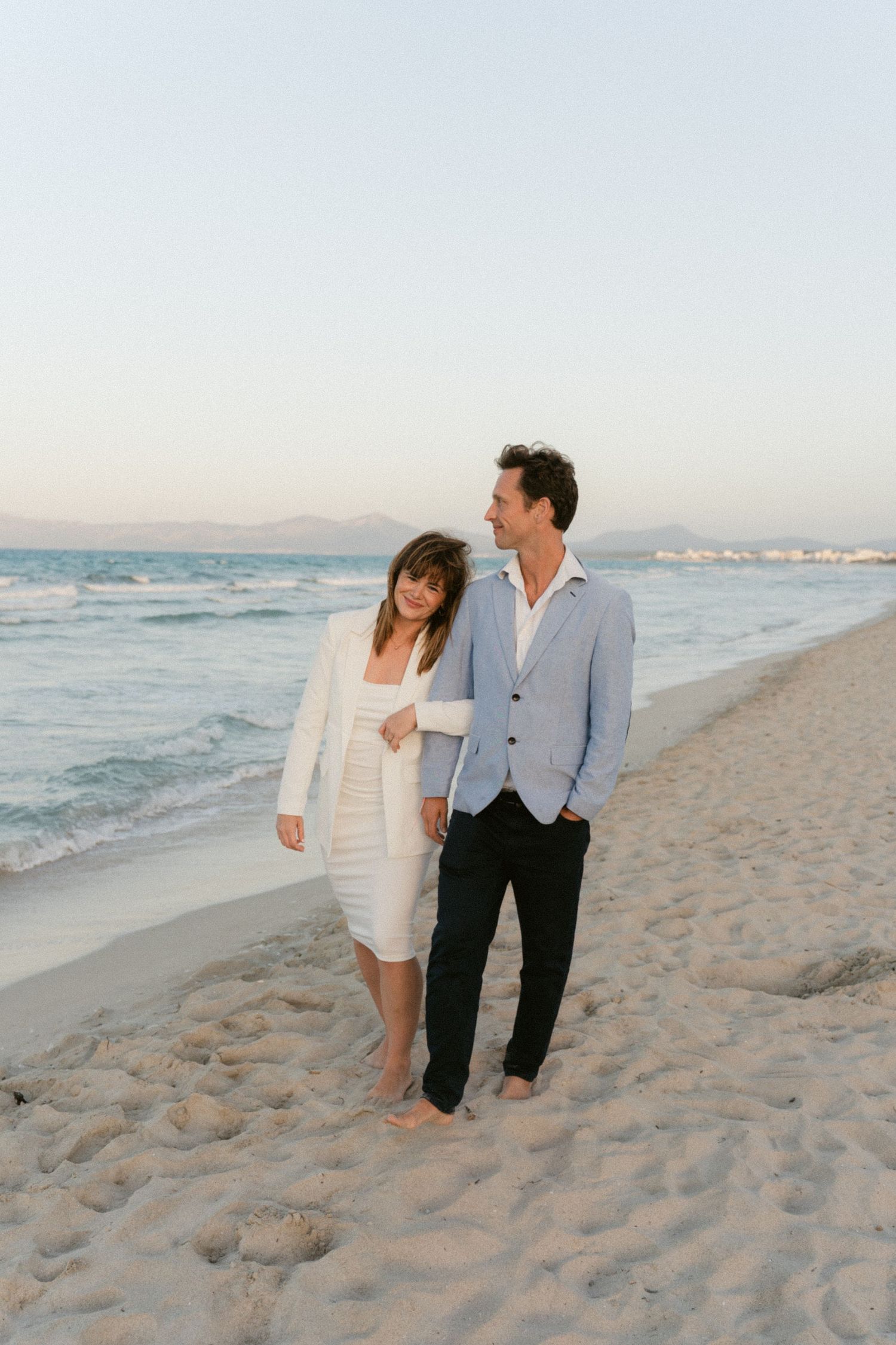 Couple in casual beach attire walking along shoreline at sunset.
