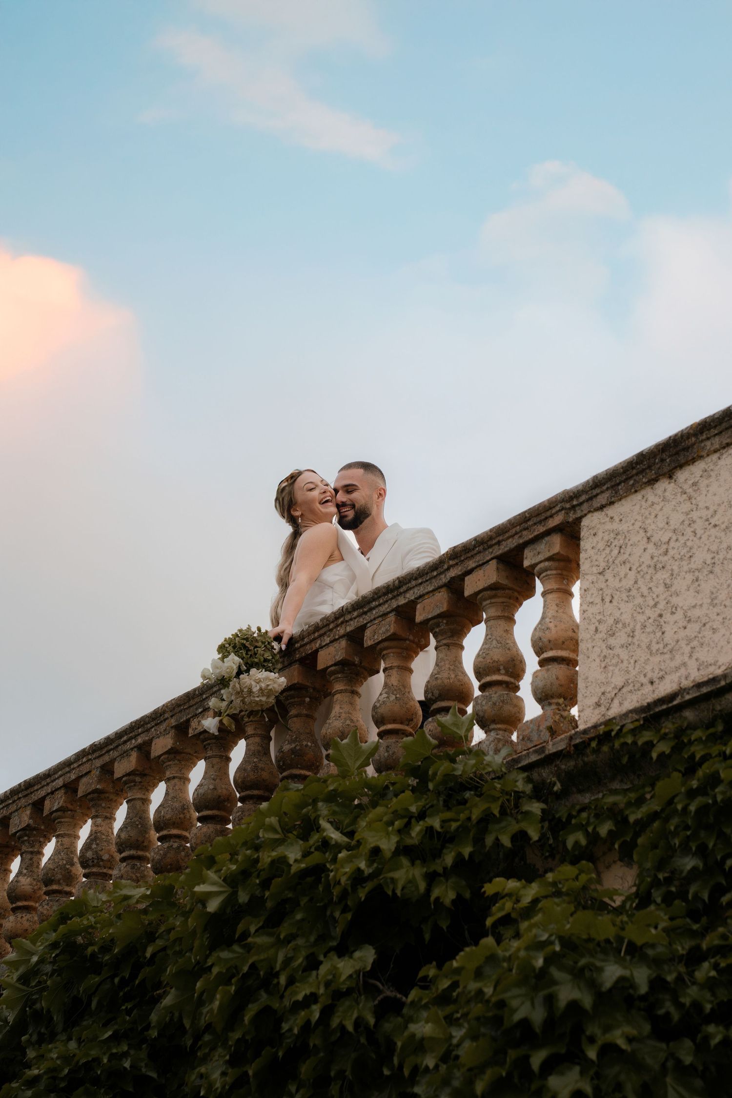 A couple shares an intimate moment along a stone balustrade against a blue sky.