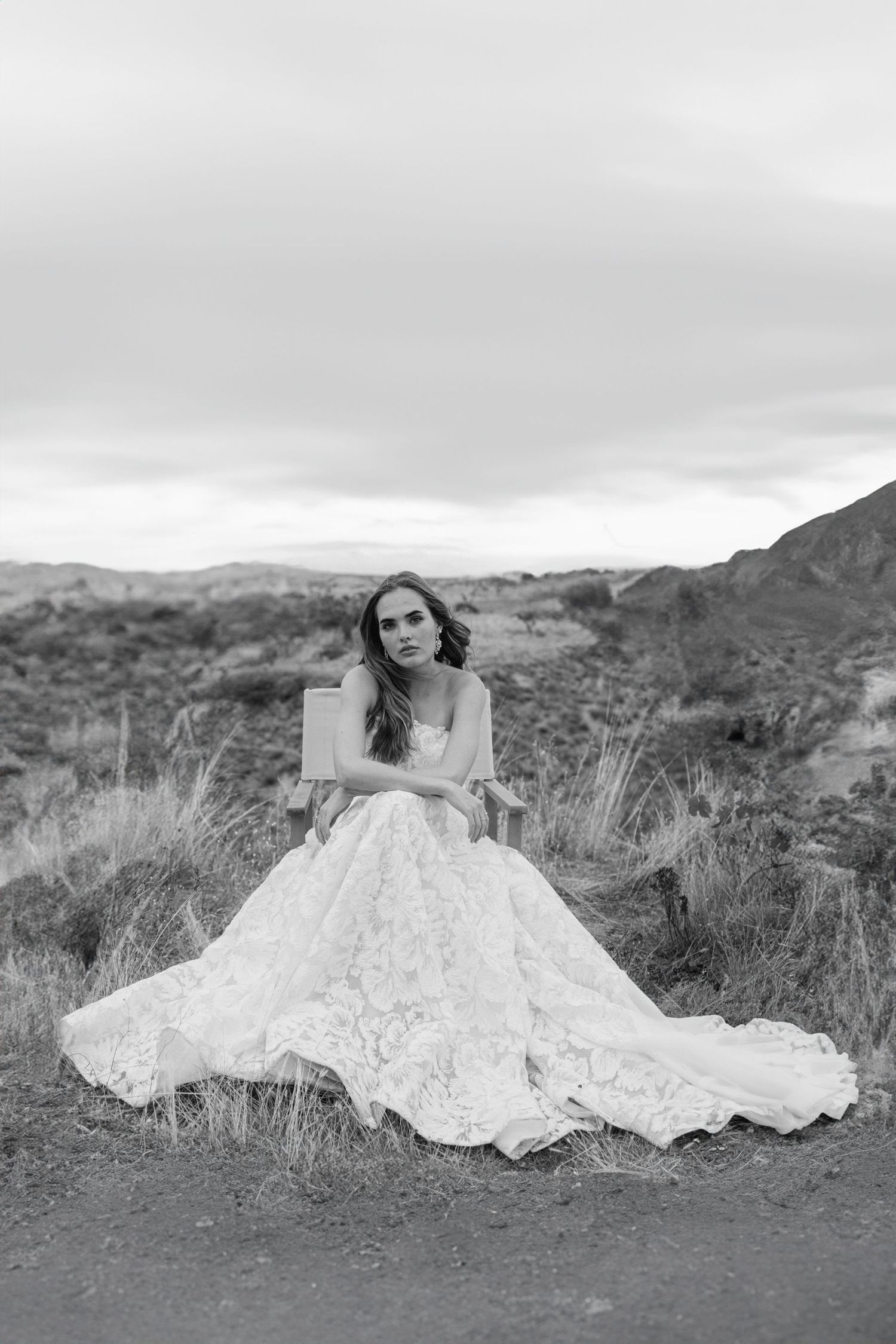A bride in a lace wedding dress poses gracefully on a grassy hillside against a mountainous backdrop.