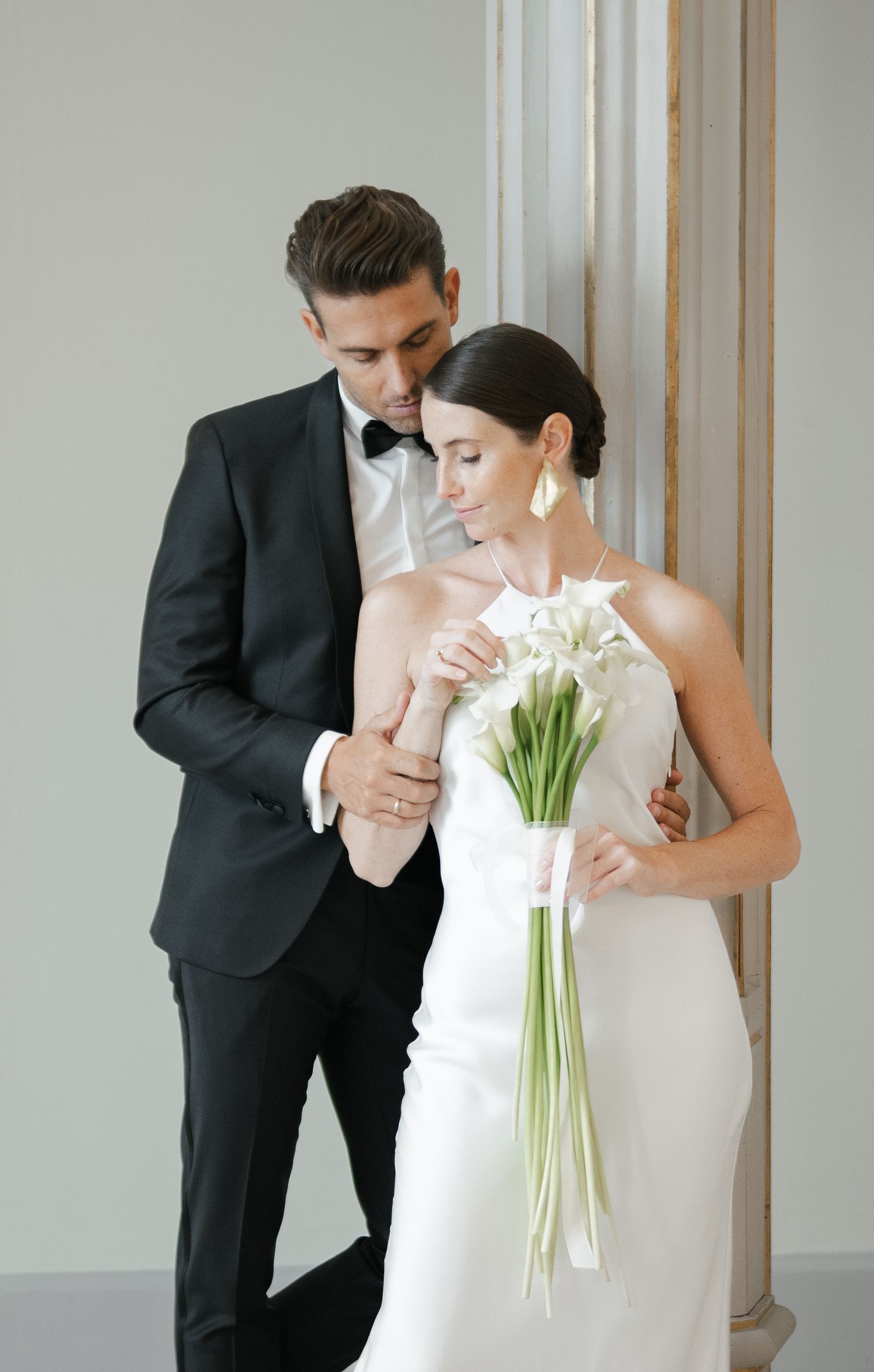 Elegant bride in a white halter gown holding a calla lily bouquet as the groom in a black tuxedo and bow tie embraces her from behind.