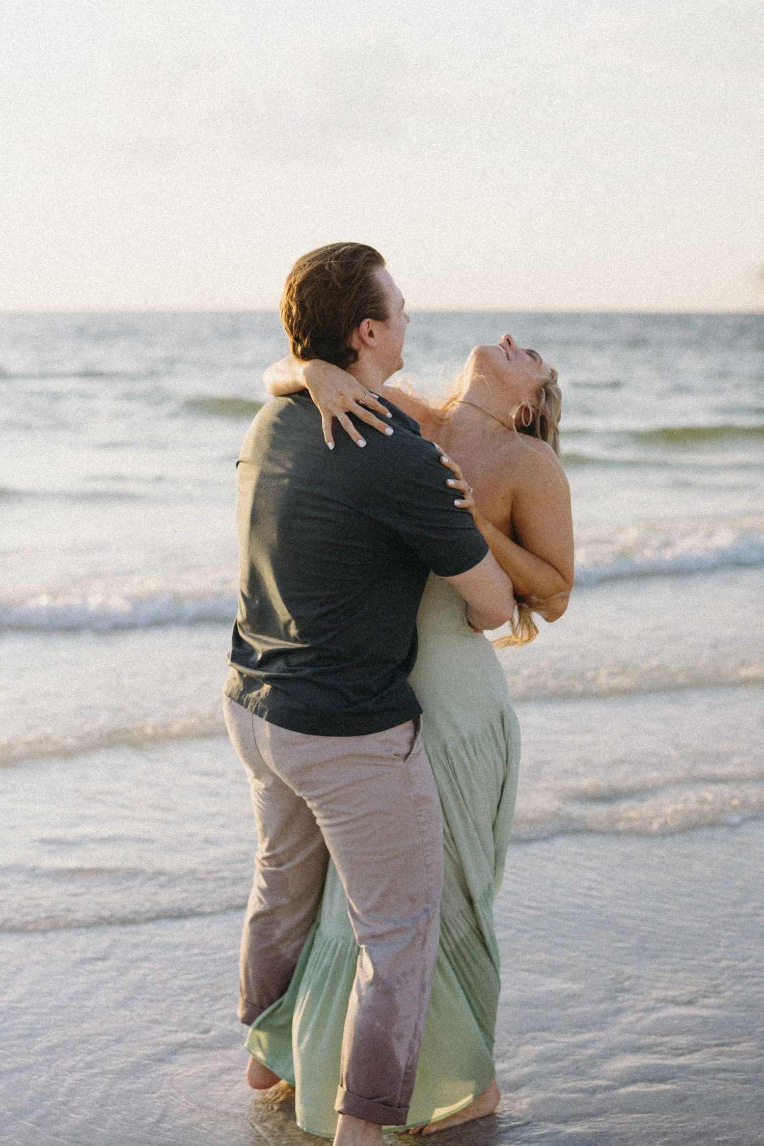 Playful couple dancing and embracing on beach with waves in background at dusk.