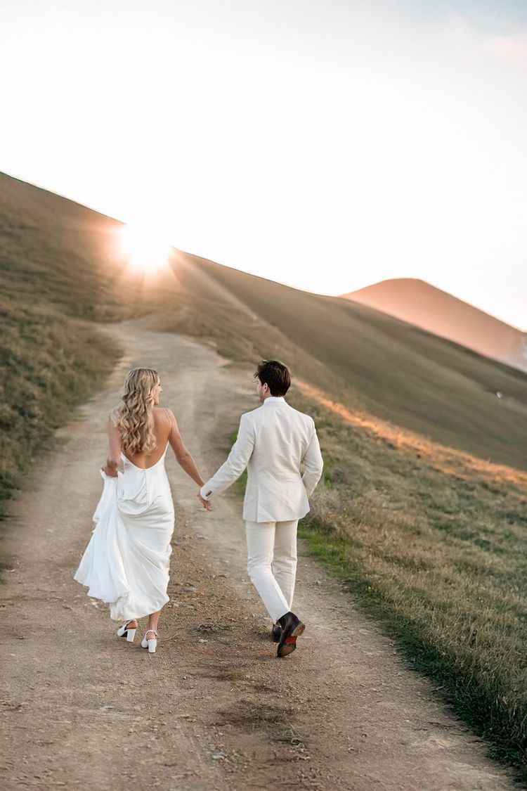 Couple in white attire running along hillside path at sunset.