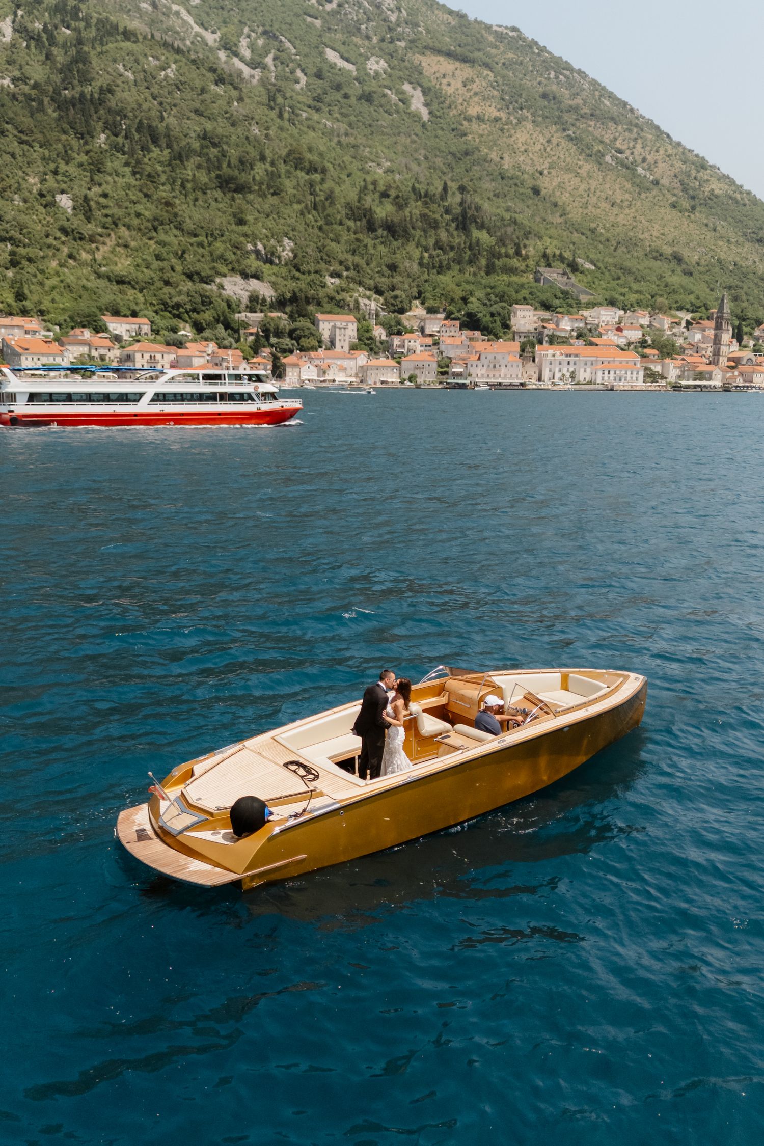 A wooden speedboat cruises across blue Mediterranean waters near a coastal mountain village.