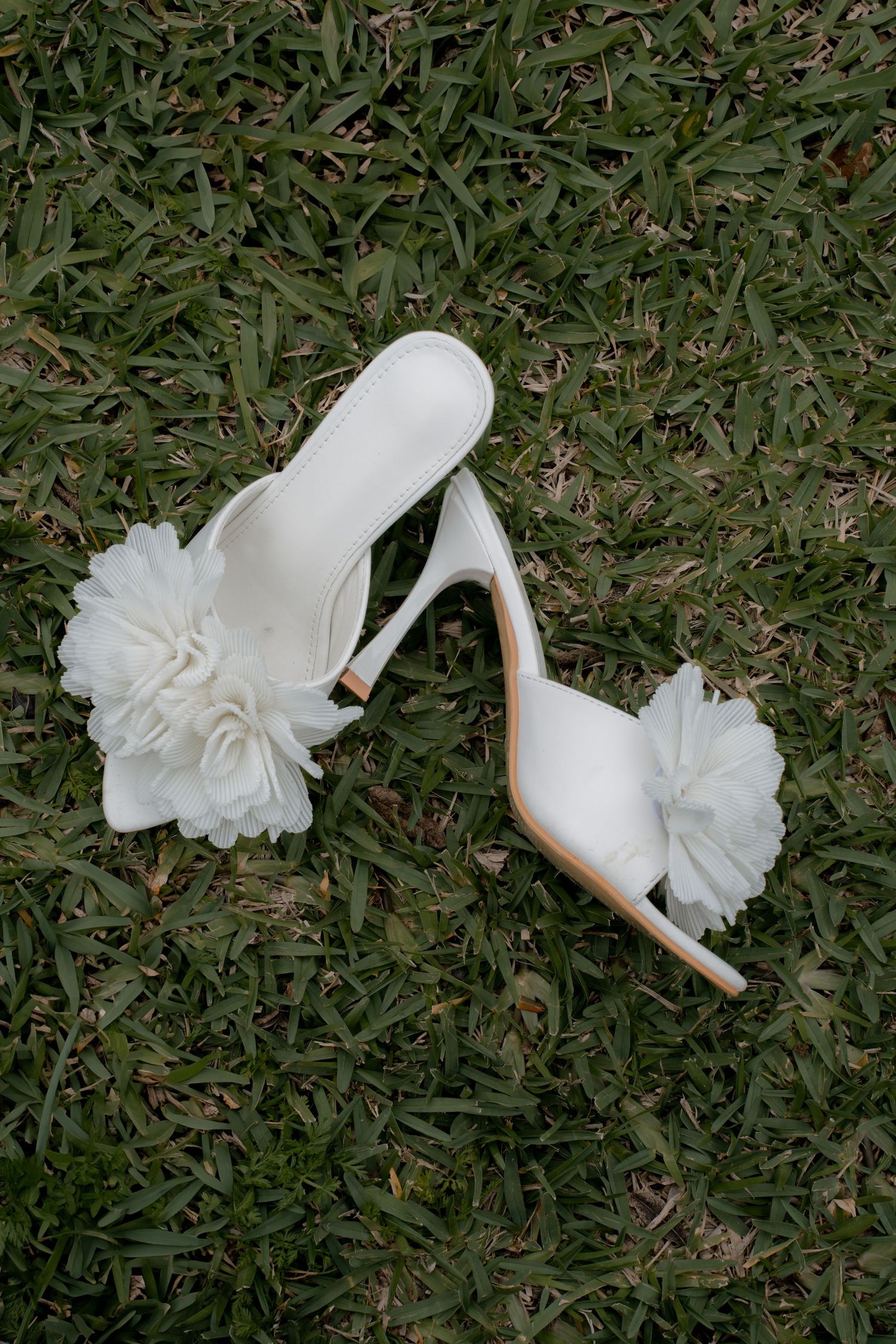 White bridal shoes with floral details lying on green grass.