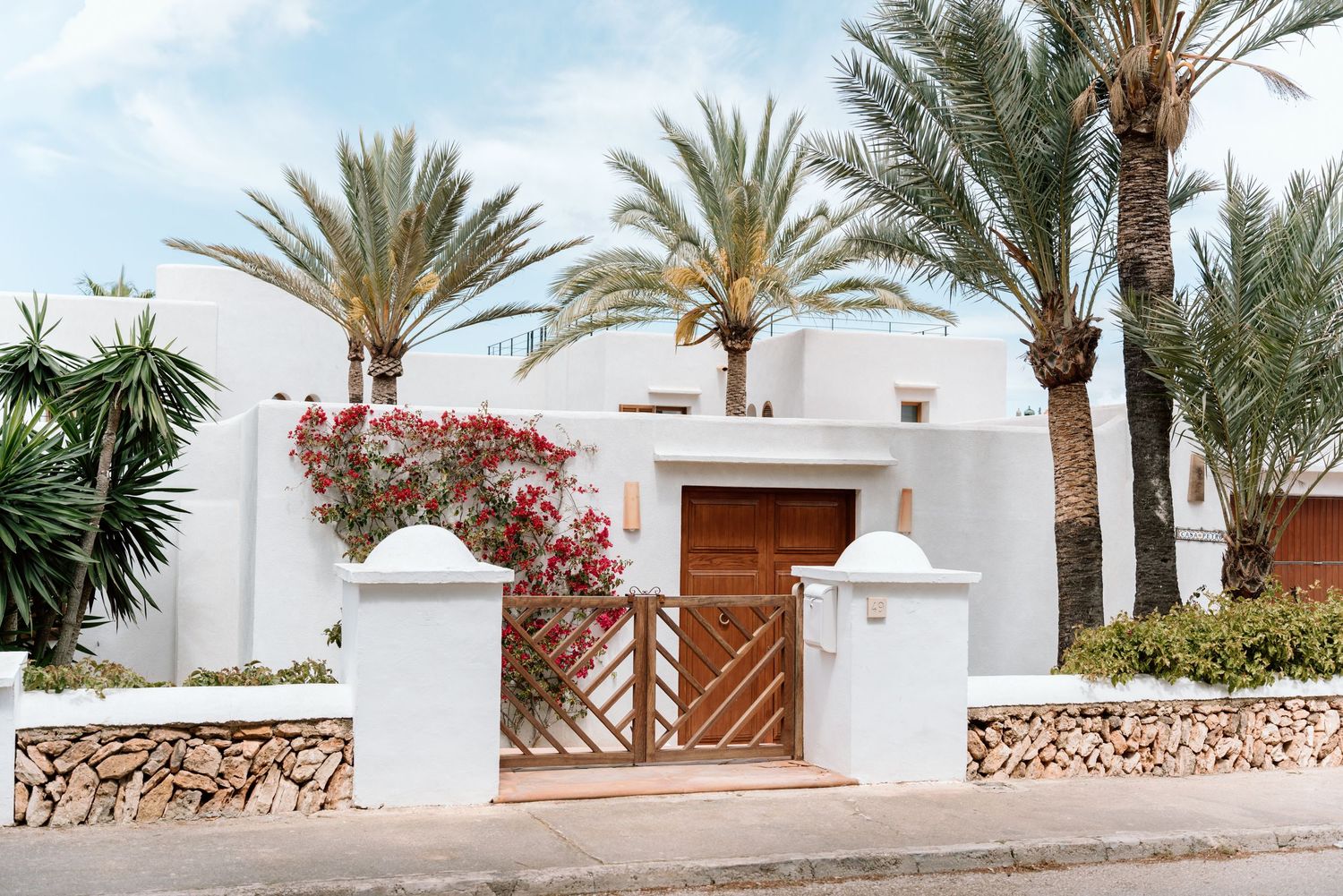 White Mediterranean villa entrance with wooden gate and palm trees along a stone pathway.