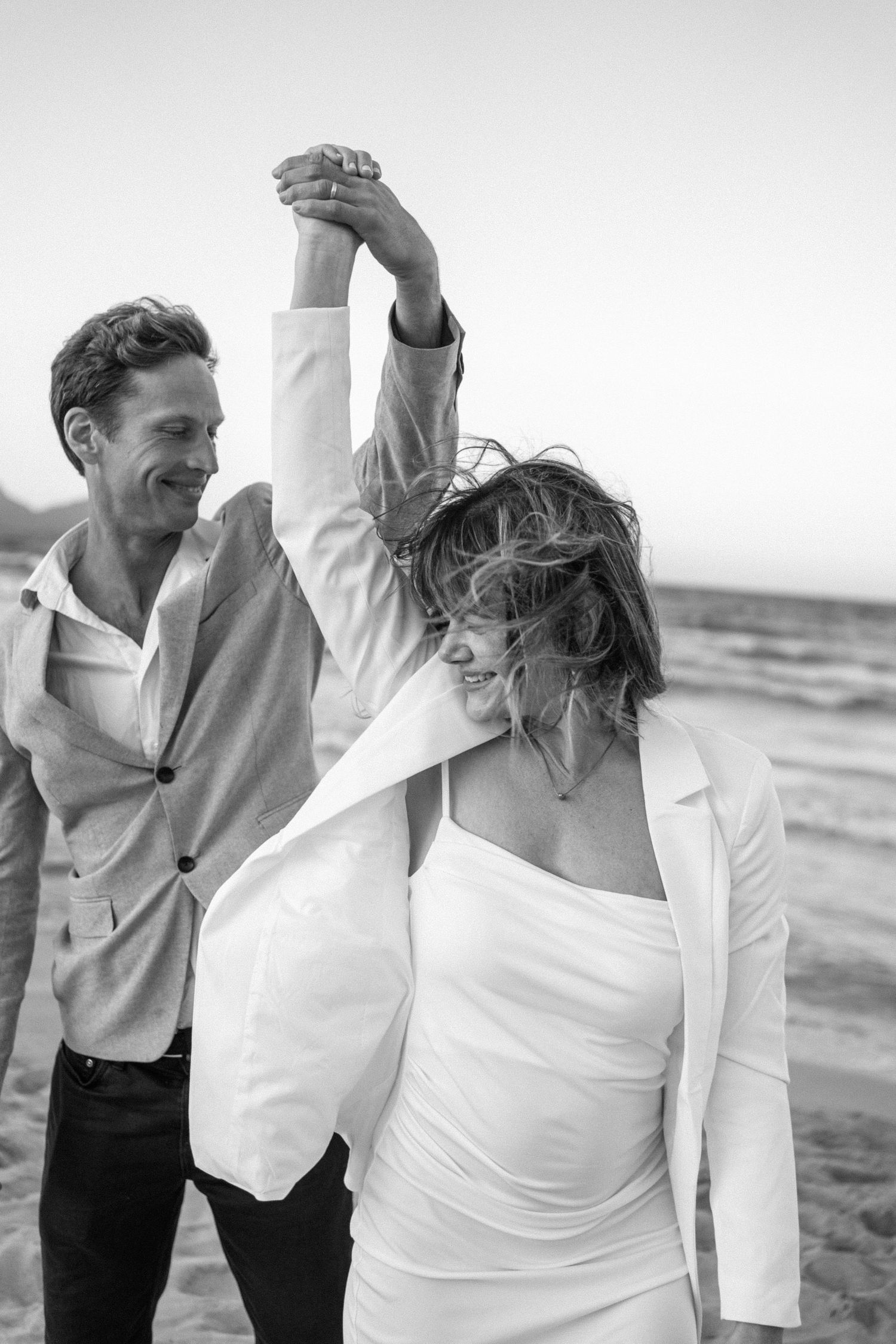 Couple in white outfits dancing joyfully on beach in black and white photography.